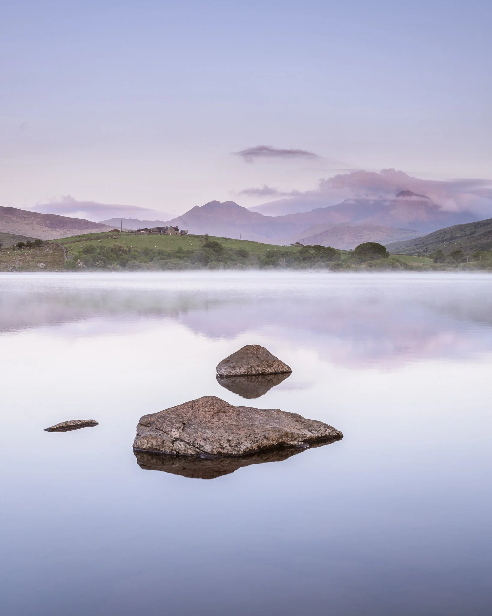 Calm lake with rocks in the foreground, mist on the water, green hills, and mountains in the background under a pastel sky.
