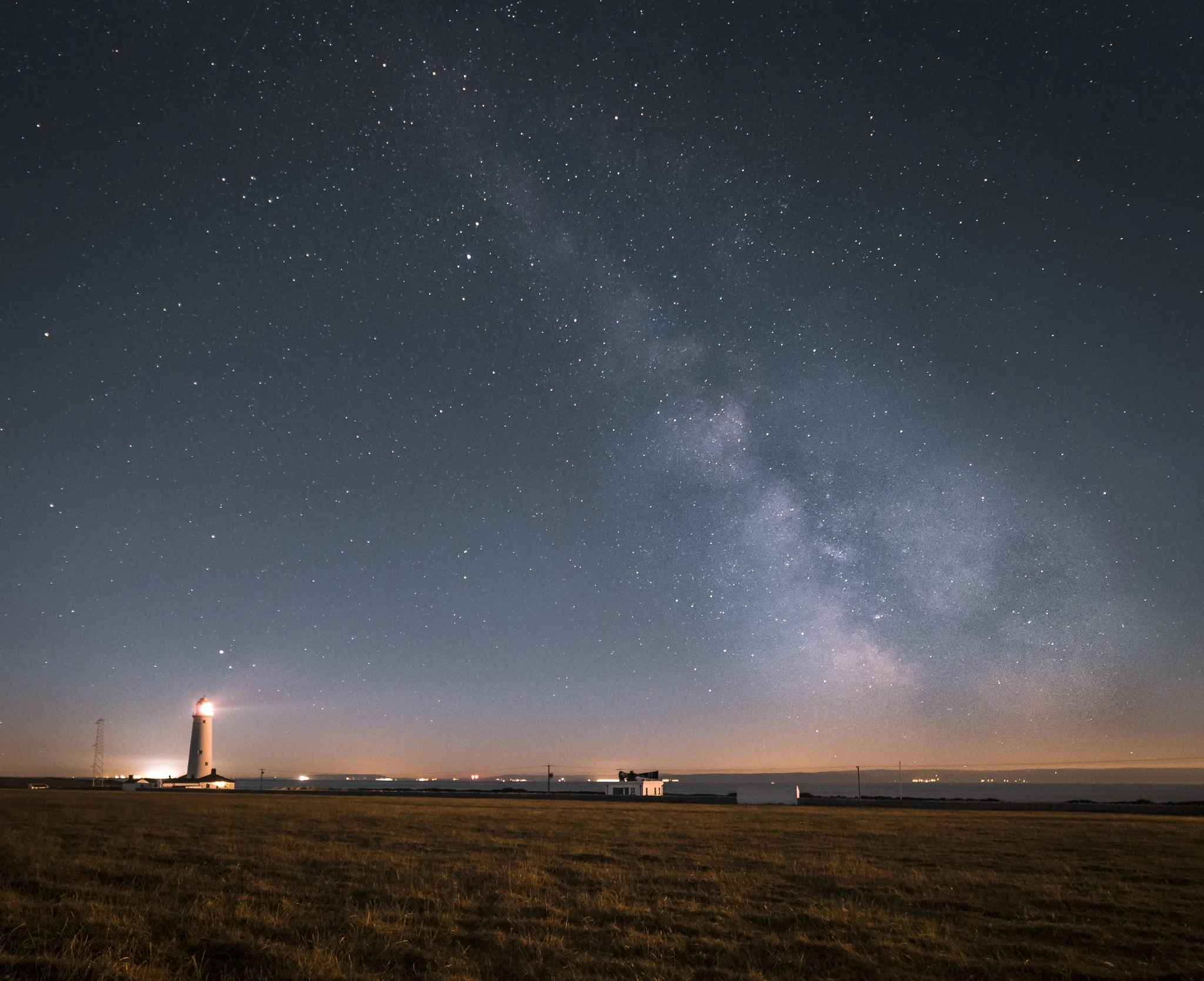 Nighttime view of the Milky Way galaxy in the sky over a rural landscape with a lighthouse, small building, and electrical poles.