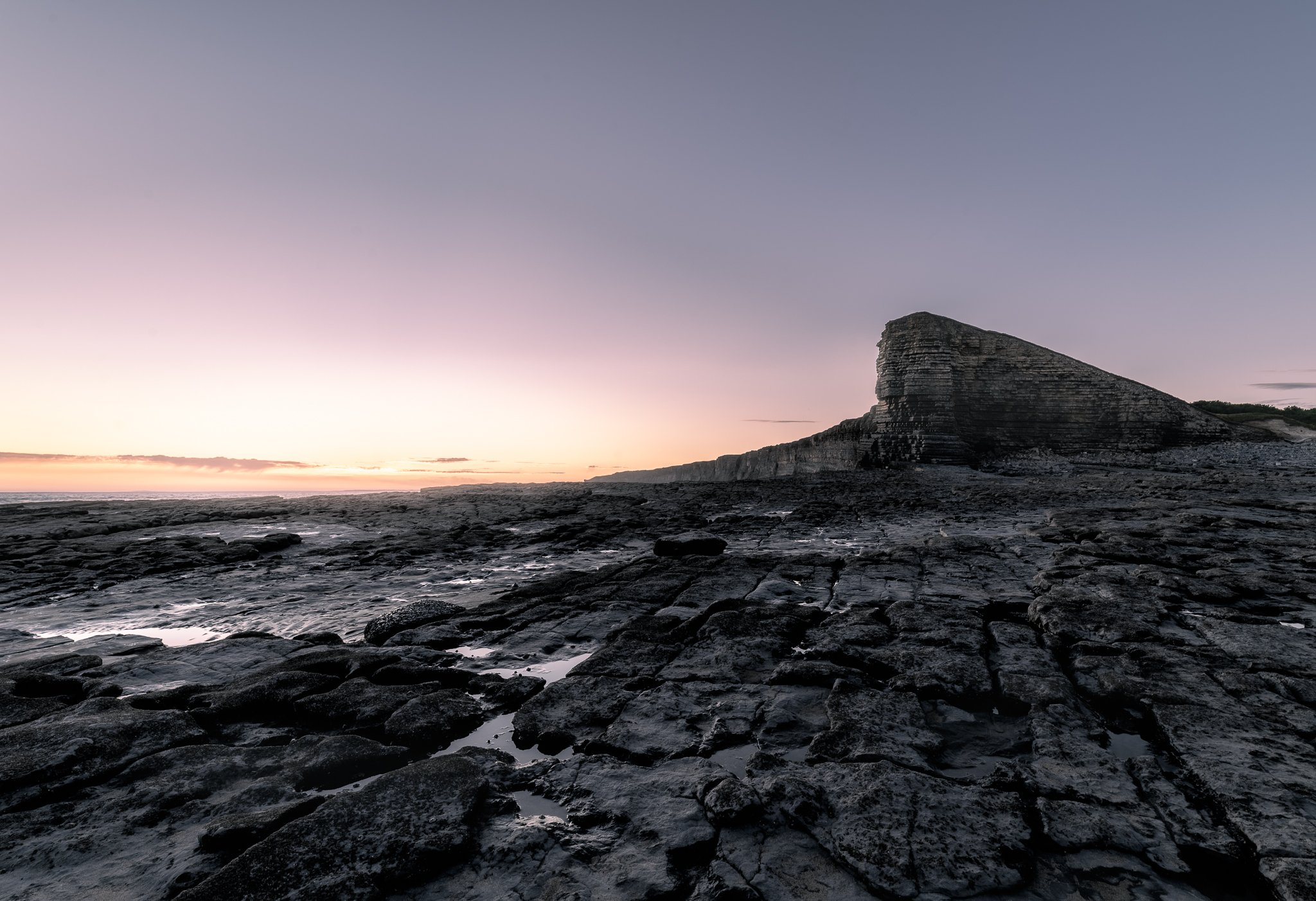 Rocky shoreline at sunset with a large layered rock formation in the distance.