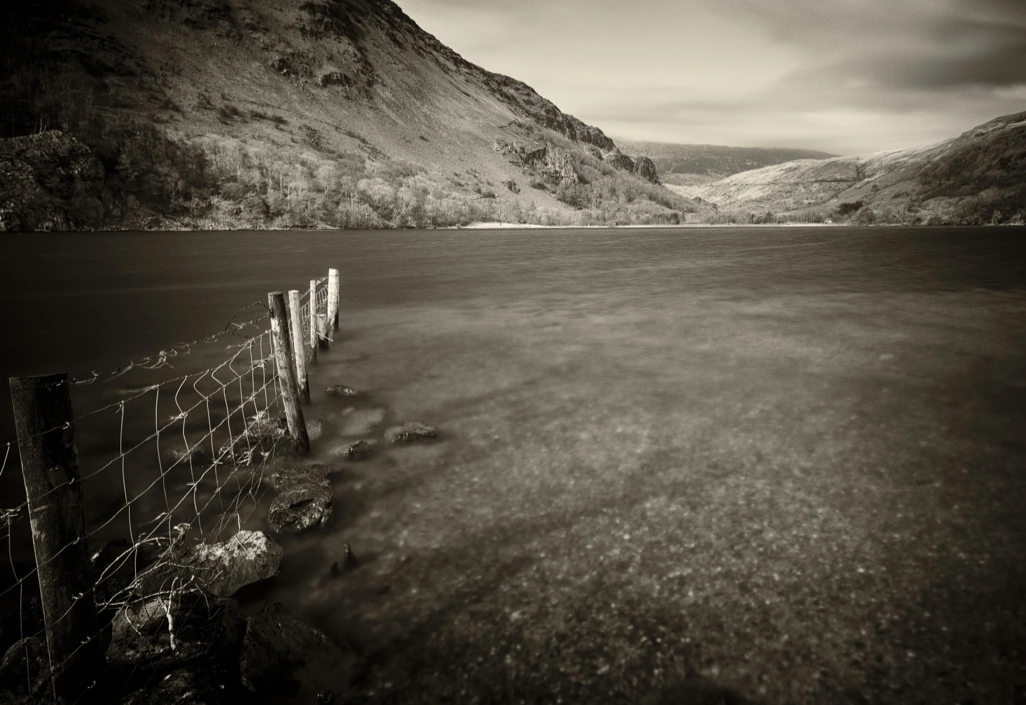 A black and white photo of a lake with a shoreline, a partially submerged fence along the water's edge, and hills or mountains in the background under a cloudy sky.