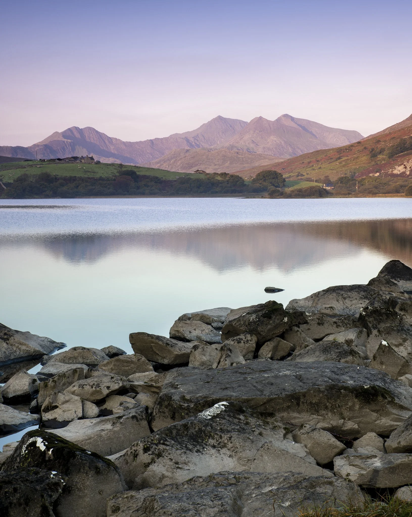 Calm lake with rocky shoreline in foreground, green hillside with trees in middle ground, and mountains in background under a pastel sky.
