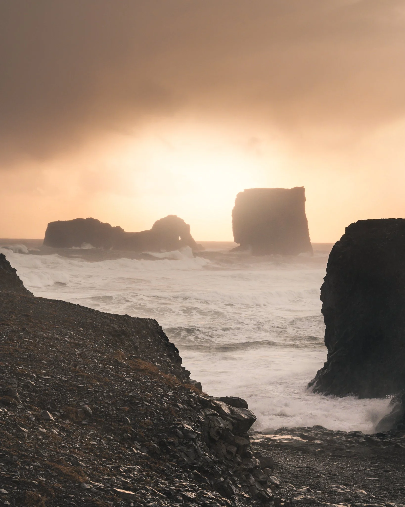 Sandy beach with dark rocks and large sea stacks during sunset.