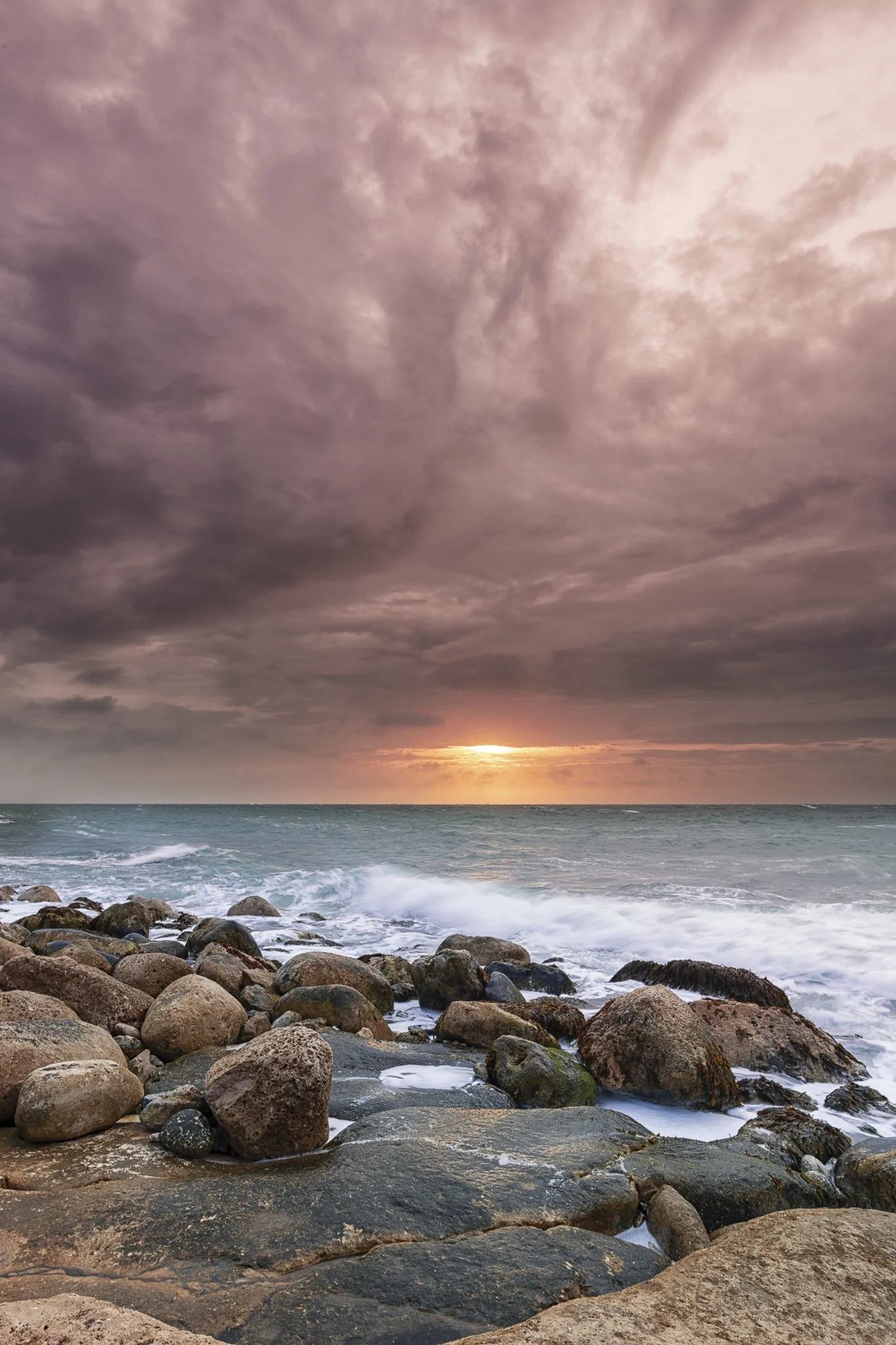 Sunset over a rocky beach with waves crashing and a cloudy sky.