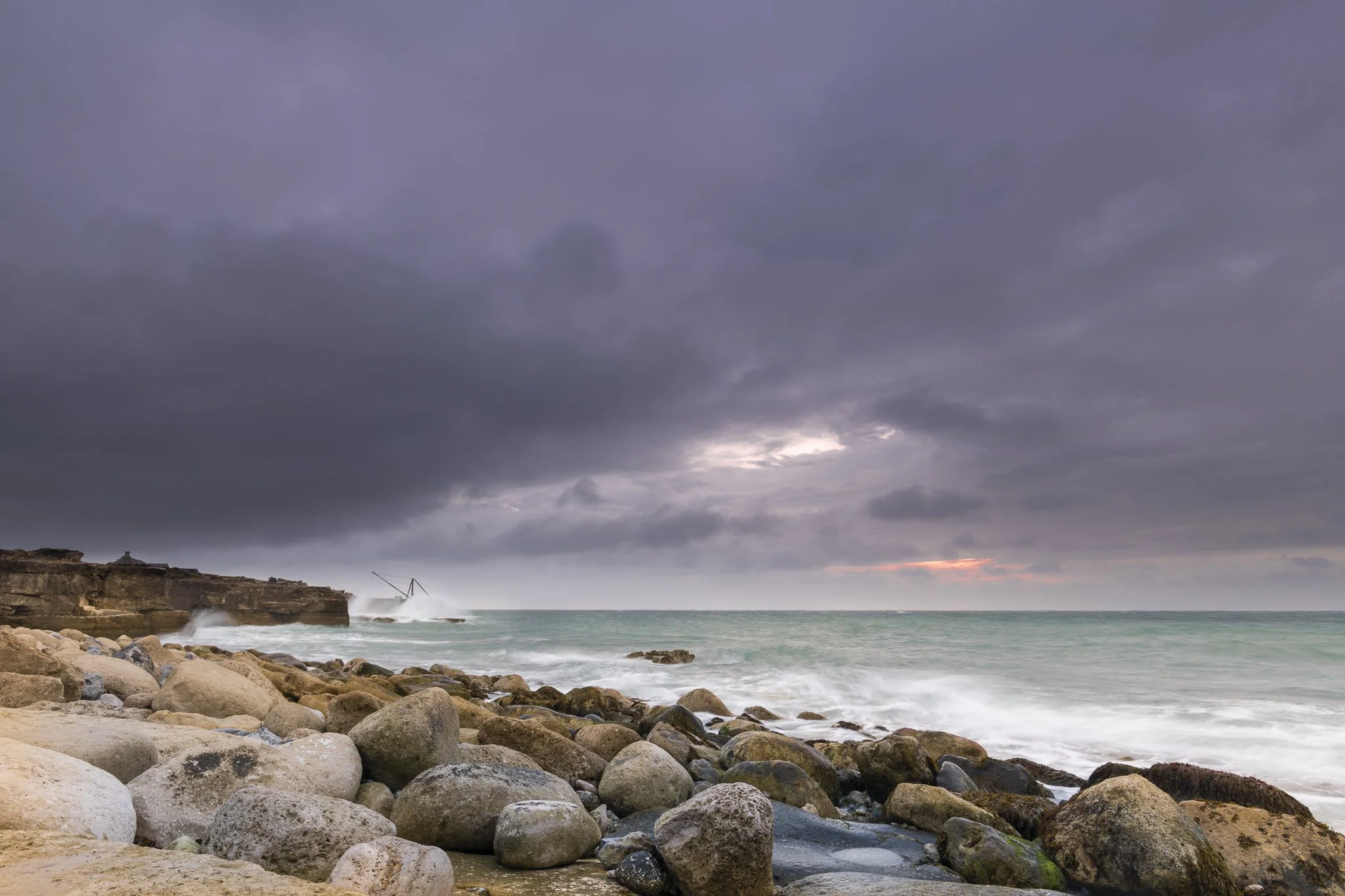 A rocky beach under a dark, cloudy sky with some orange hues near the horizon, ocean waves crashing against the rocks, and a leaning structure or pole in the distance on the left side.