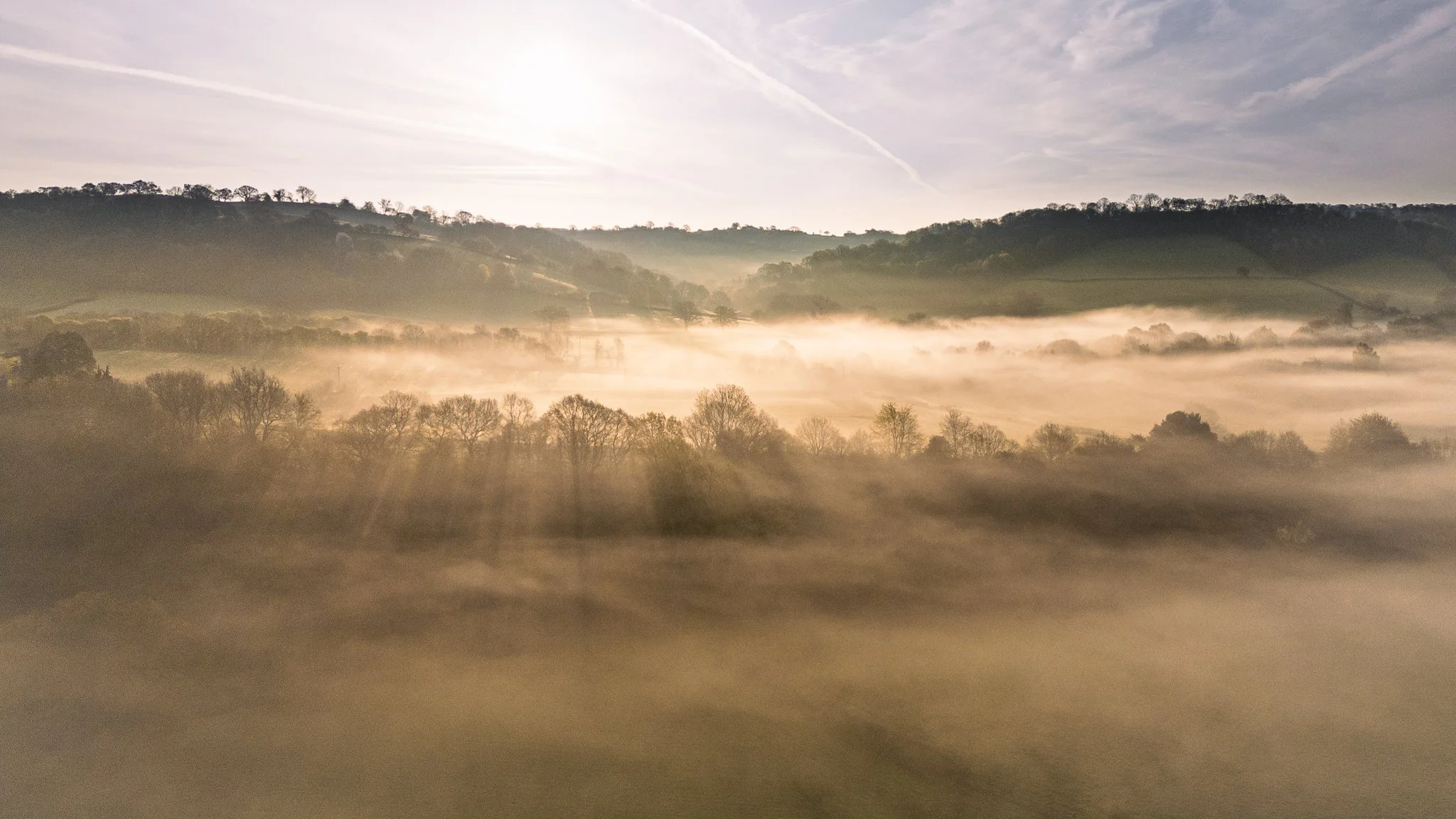 A scenic landscape of rolling hills and trees during sunrise or sunset with mist or fog covering the land and rays of sunlight shining through.