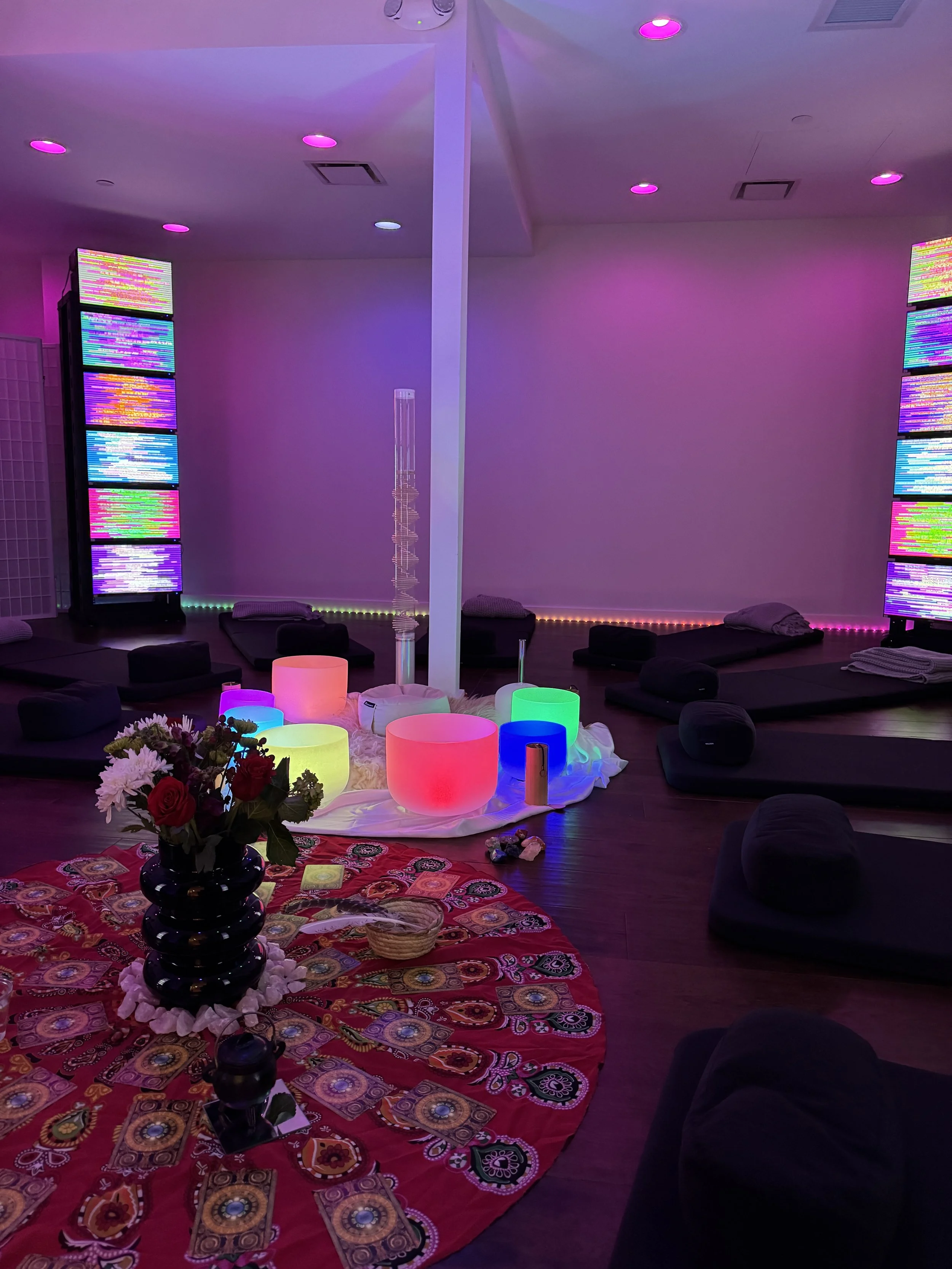A room with colorful LED lights, black cushions on the floor, a circular red cloth with a black vase of flowers, glowing rounded objects, and decorative light displays.