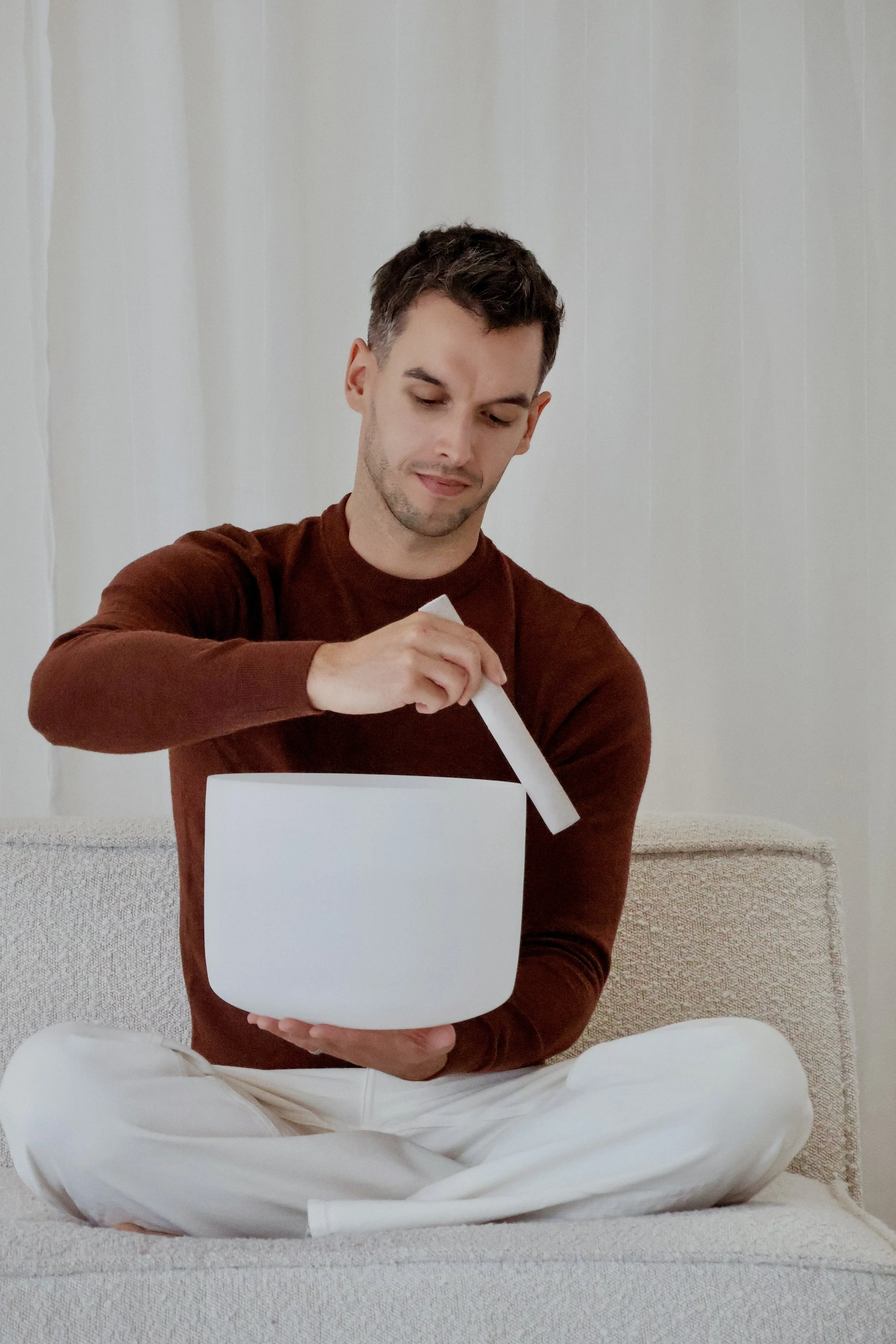 A young man sitting, holding a white singing bowl in one hand and a white mallet in the other, preparing to play.