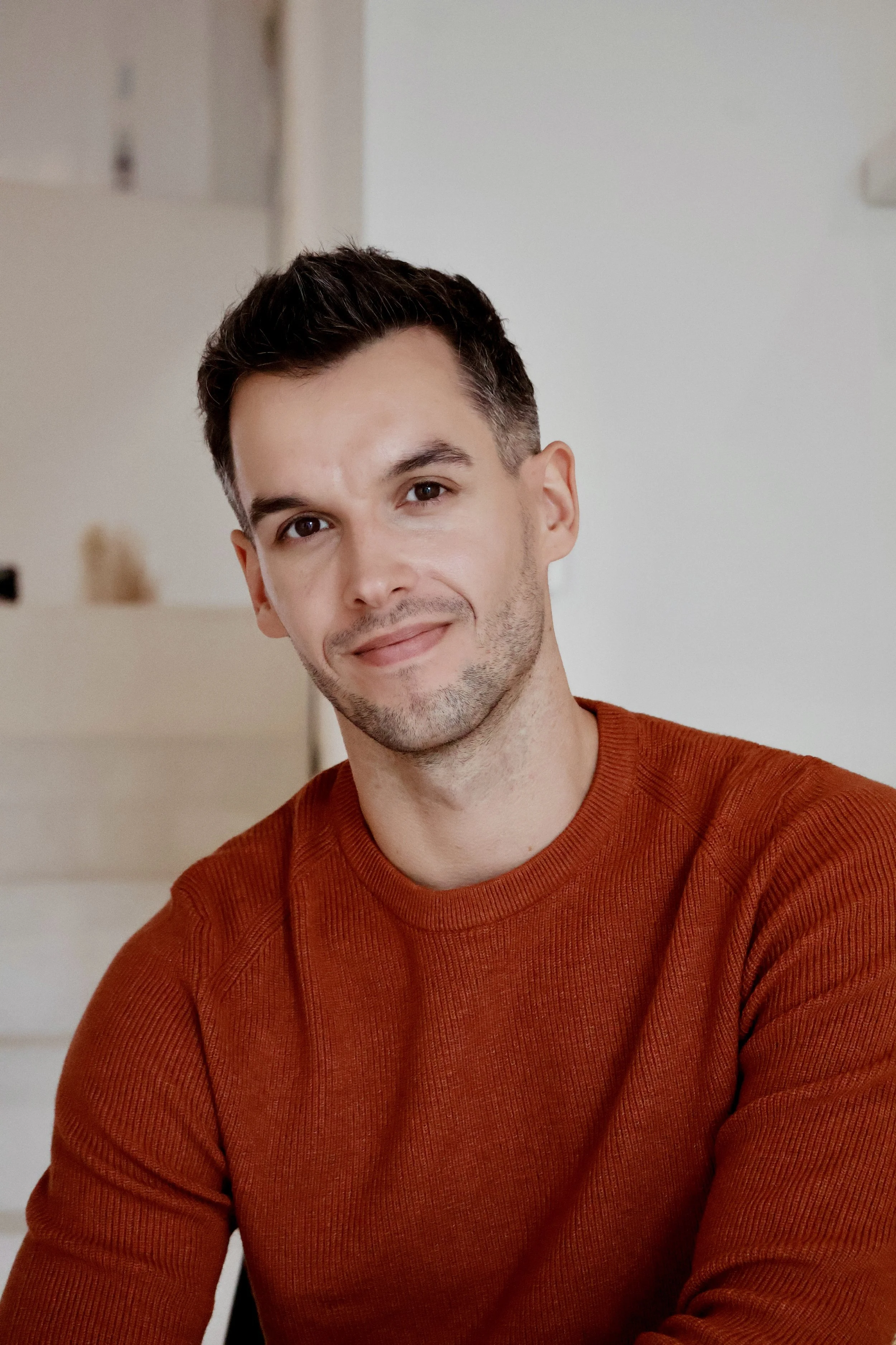 A smiling man with dark hair and facial stubble, wearing an orange sweater, sitting indoors.
