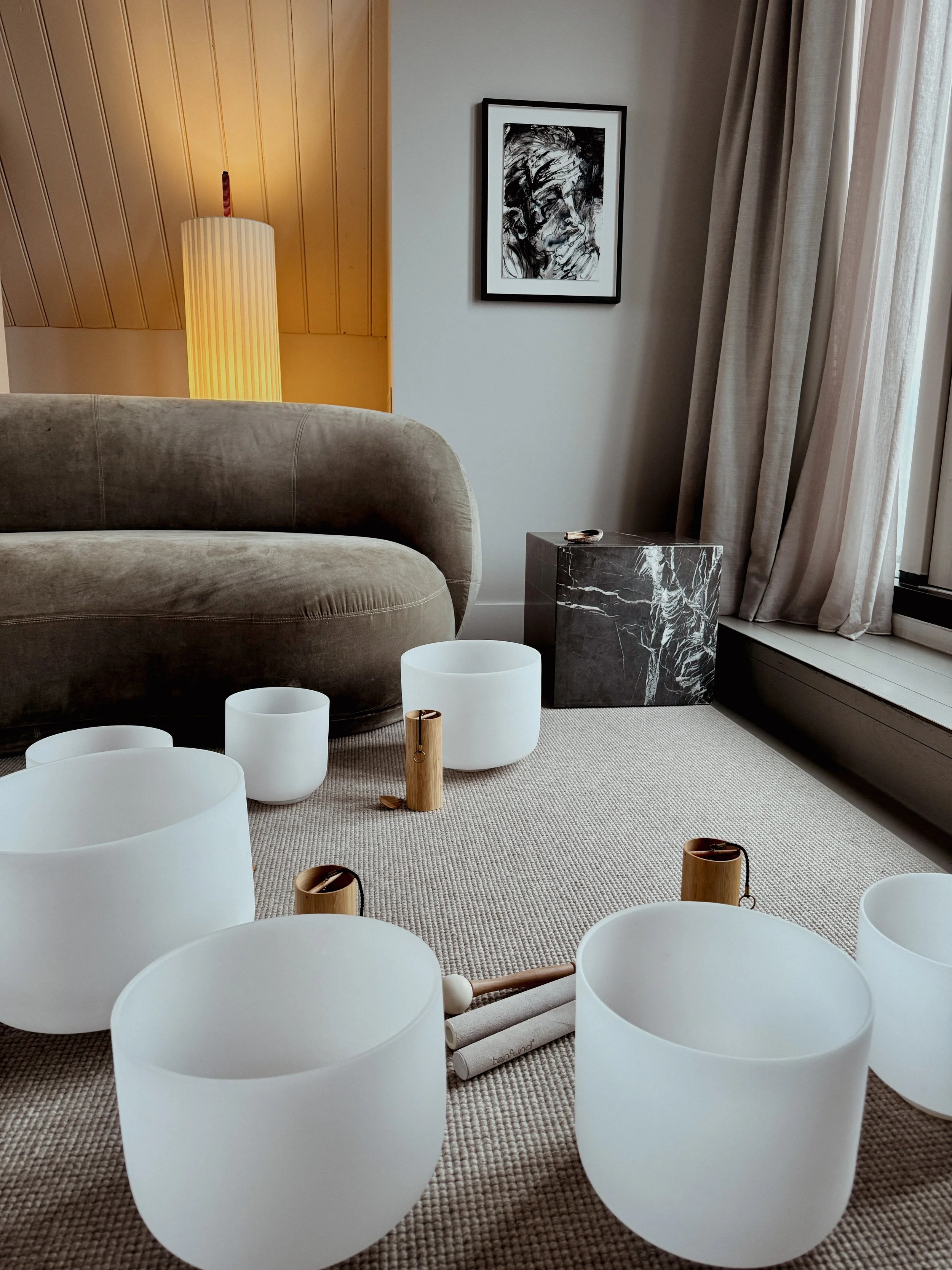 Living room with a beige sofa, a yellow floor lamp, framed black and white portrait on white wall, curtains by window, and a collection of white crystal singing bowls with wooden mallets on beige carpet.