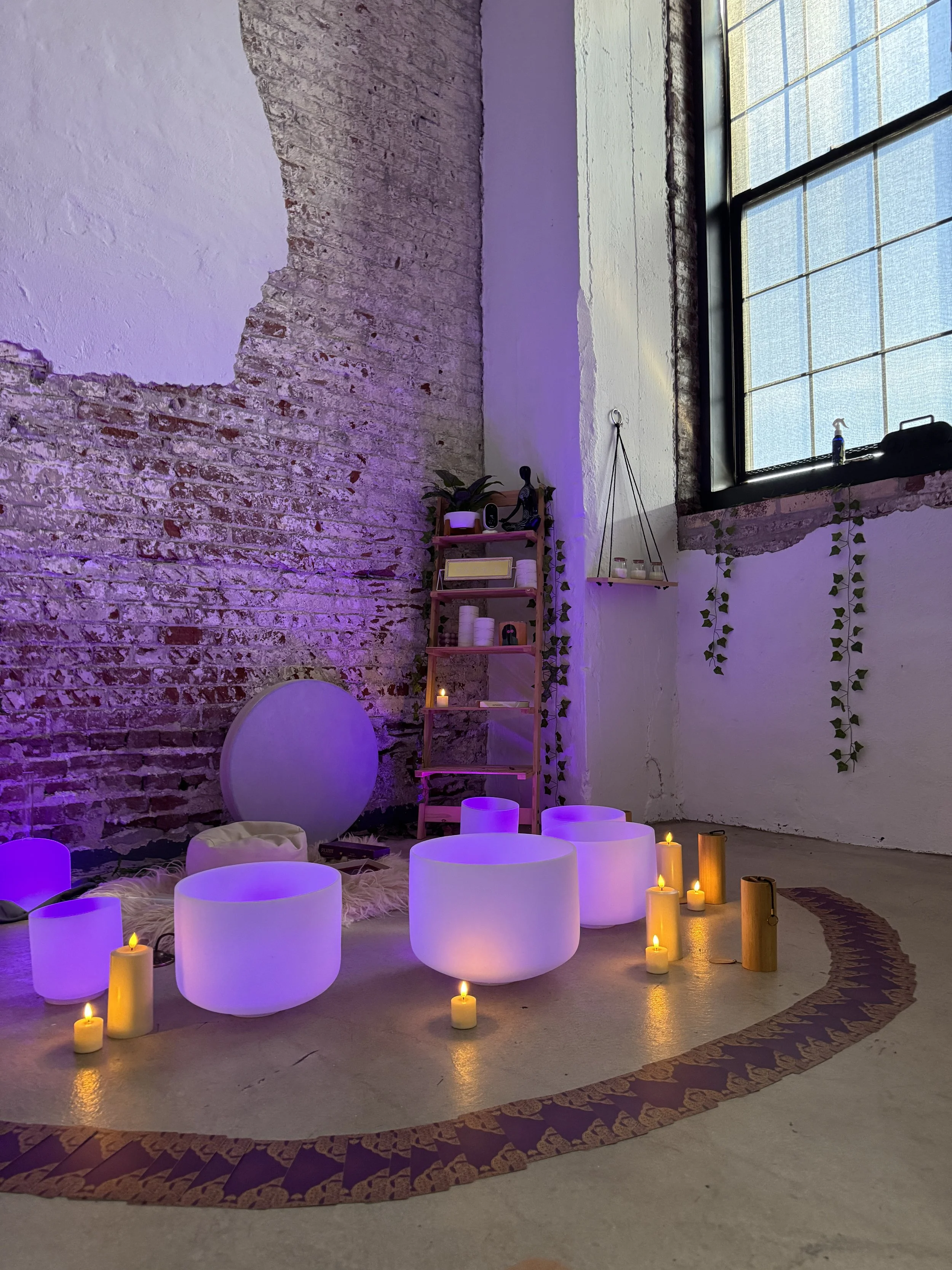 A cozy space with glowing white crystal sound bowls, various candles, a small wooden cone, a fluffy white rug, and a shelving unit with plants, books, and decorative items against a brick wall with a partially whitewashed section and a large window with hanging ivy.