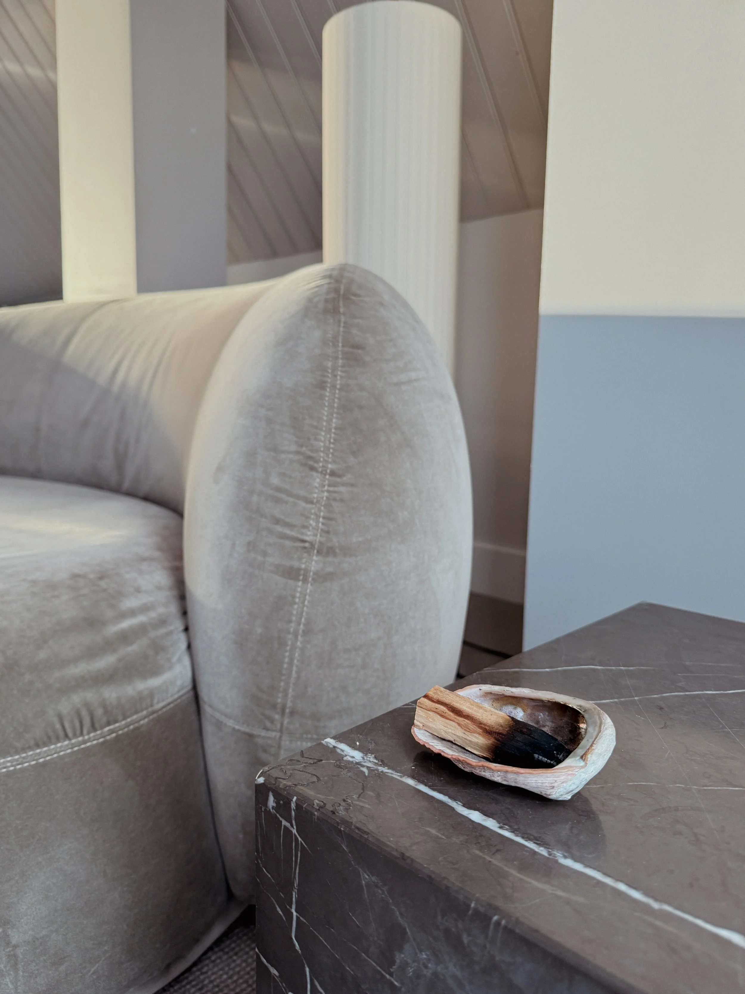 Close-up of a beige velvet sofa with a curved armrest and a dark marble side table with a small, blackened and burnt piece of palo santo wood in a decorative dish.