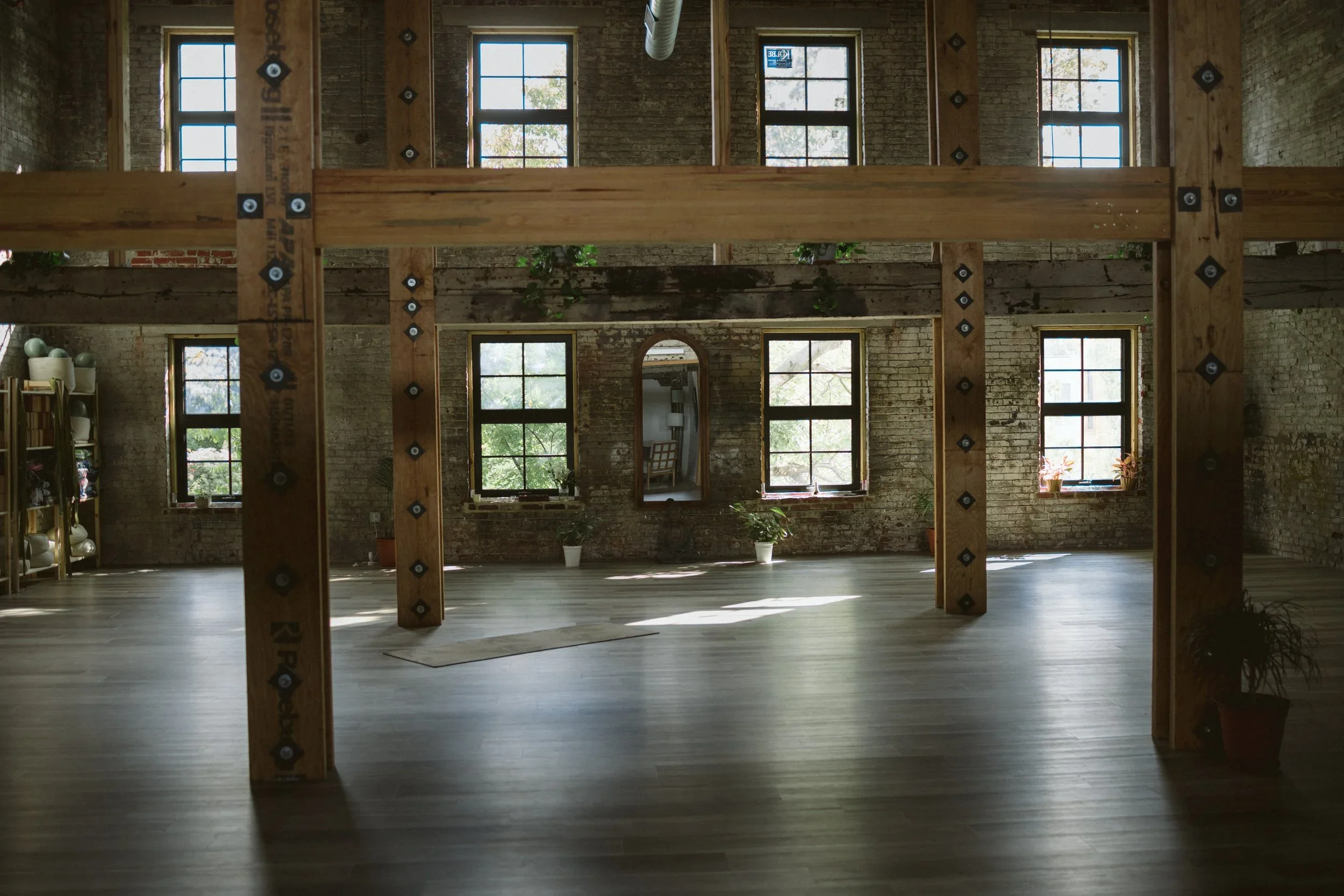 Interior view of an industrial-style space with exposed brick walls, wooden beams, multiple windows, and potted plants.