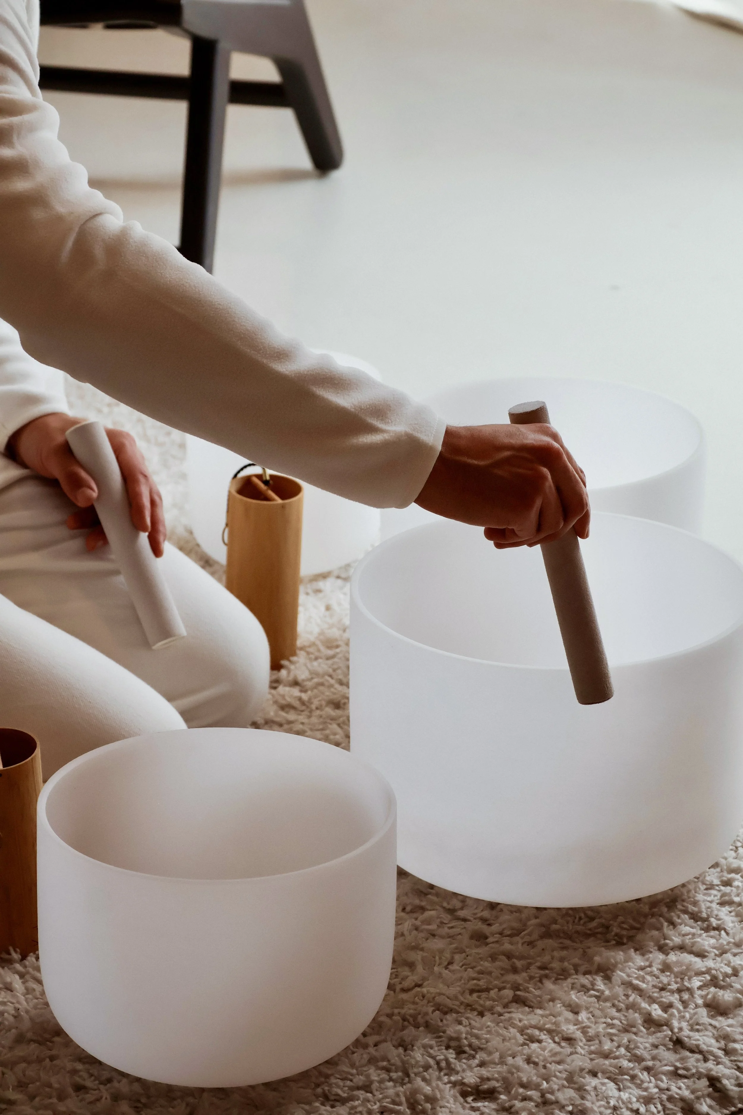 A person playing a white crystal singing bowl with a mallet, surrounded by other similar bowls on a soft beige carpet.