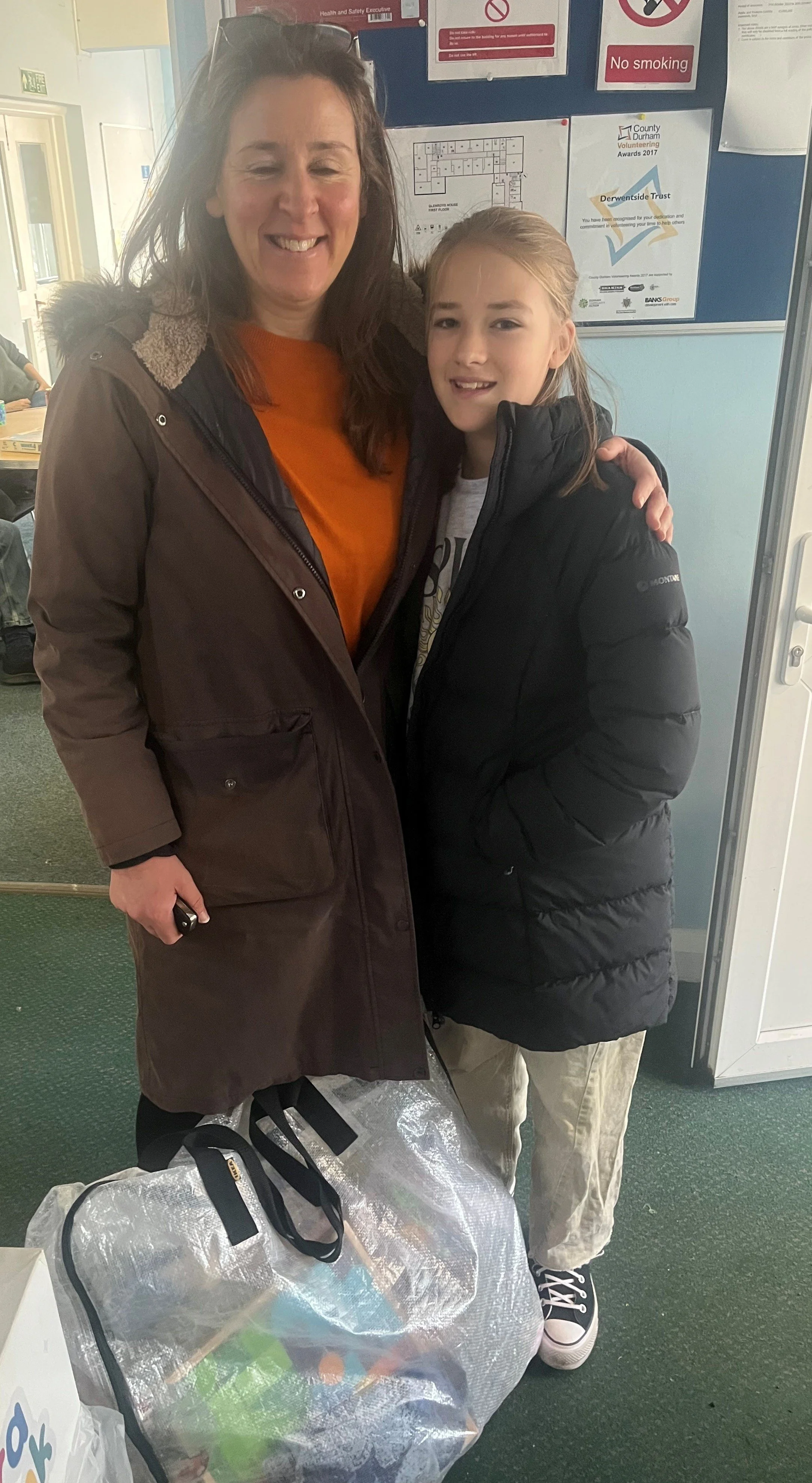 A woman and a young girl smiling and standing together indoors. The woman is wearing a brown coat and the girl is wearing a black puffer jacket. There is a large, shiny bag in front of them and a bulletin board with notices behind them.