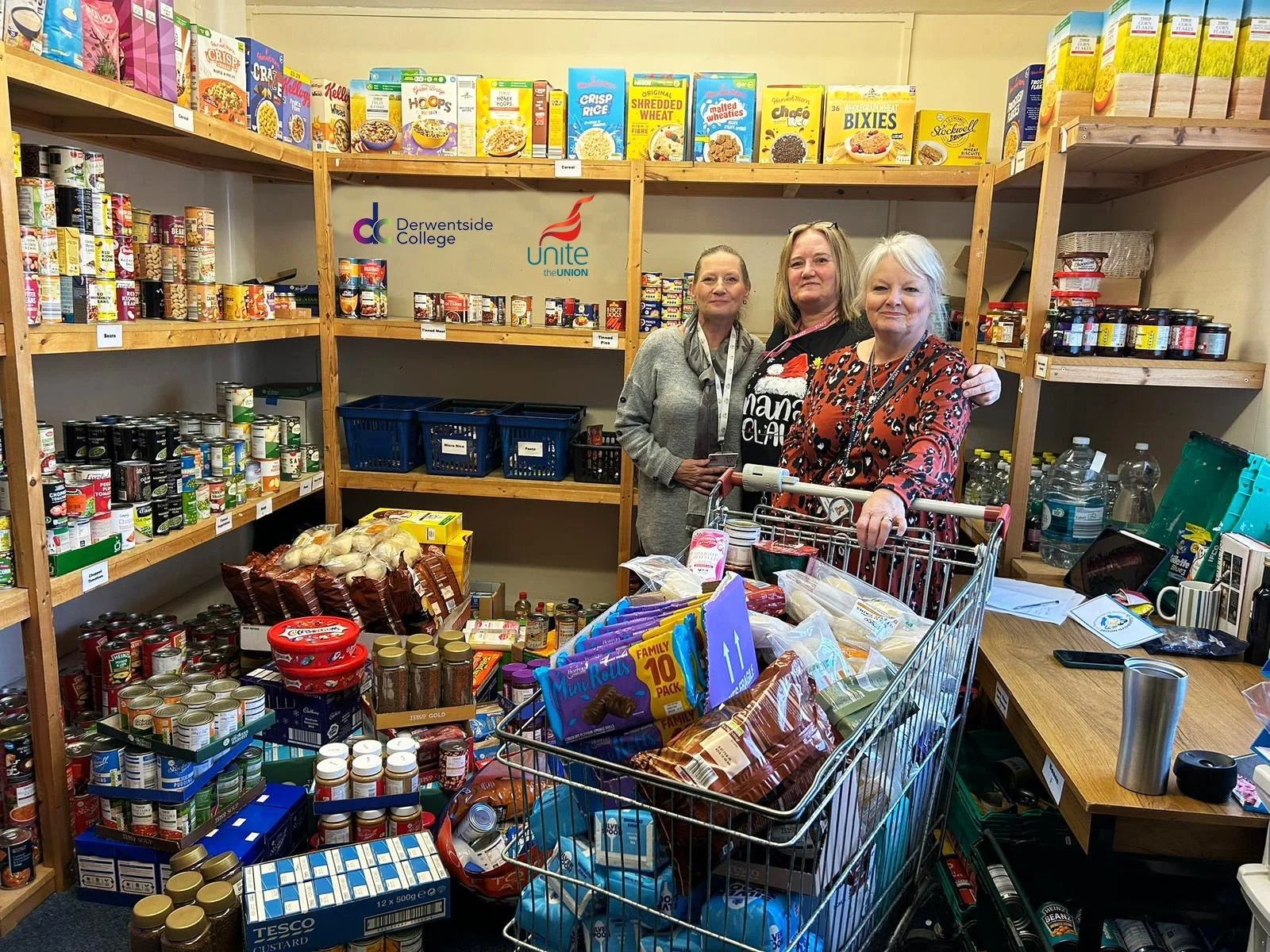 Three women standing behind a shopping cart filled with food and supplies in a food bank or donation setting, with shelves of canned goods and boxed food behind them.