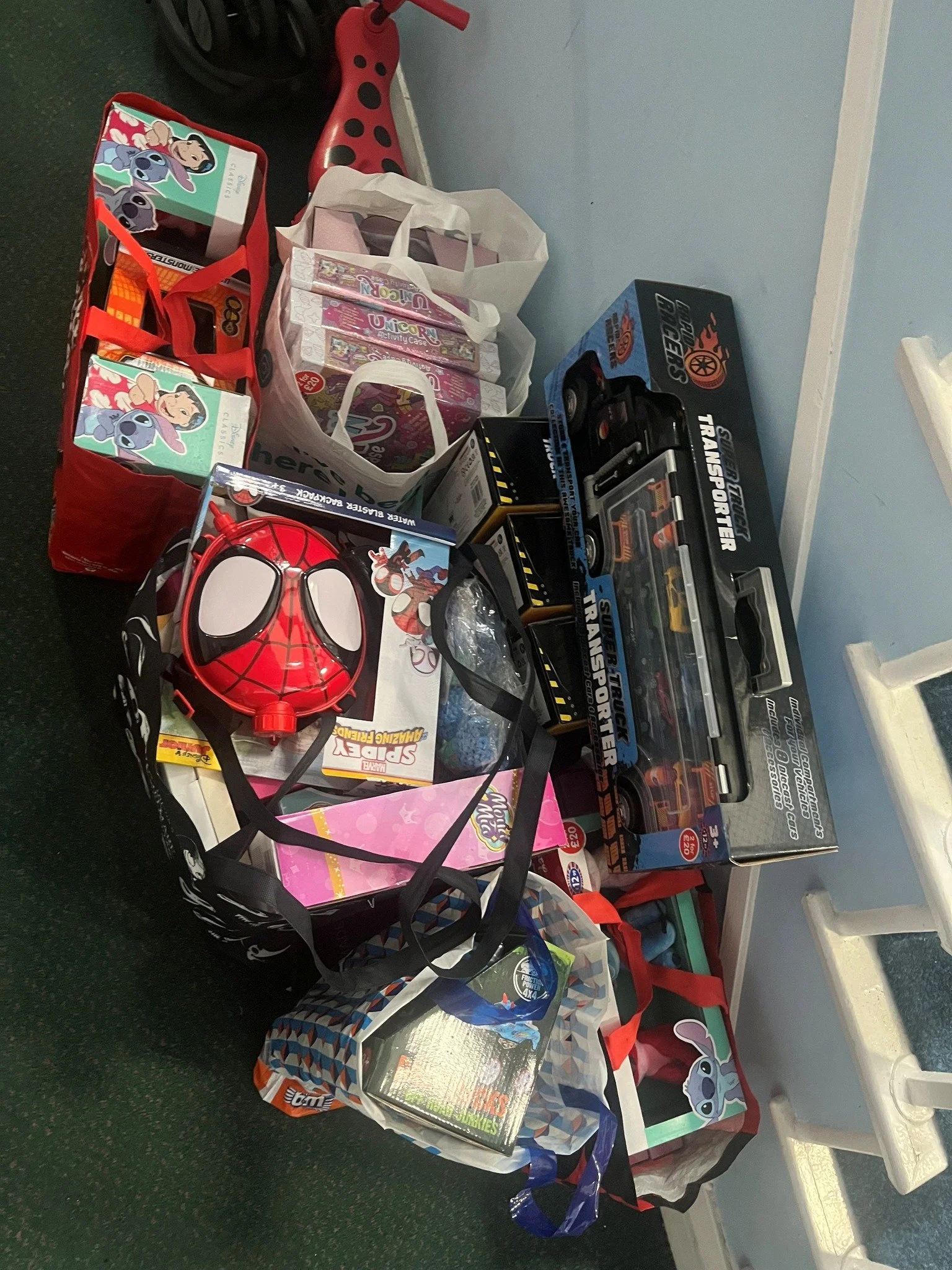 Collection of toys and gifts on the floor, including a Spider-Man mask, toy cars in boxes, a Sonic the Hedgehog plush, and a pink My Little Pony box, with shopping bags and a ladybug-themed stroller in the background.