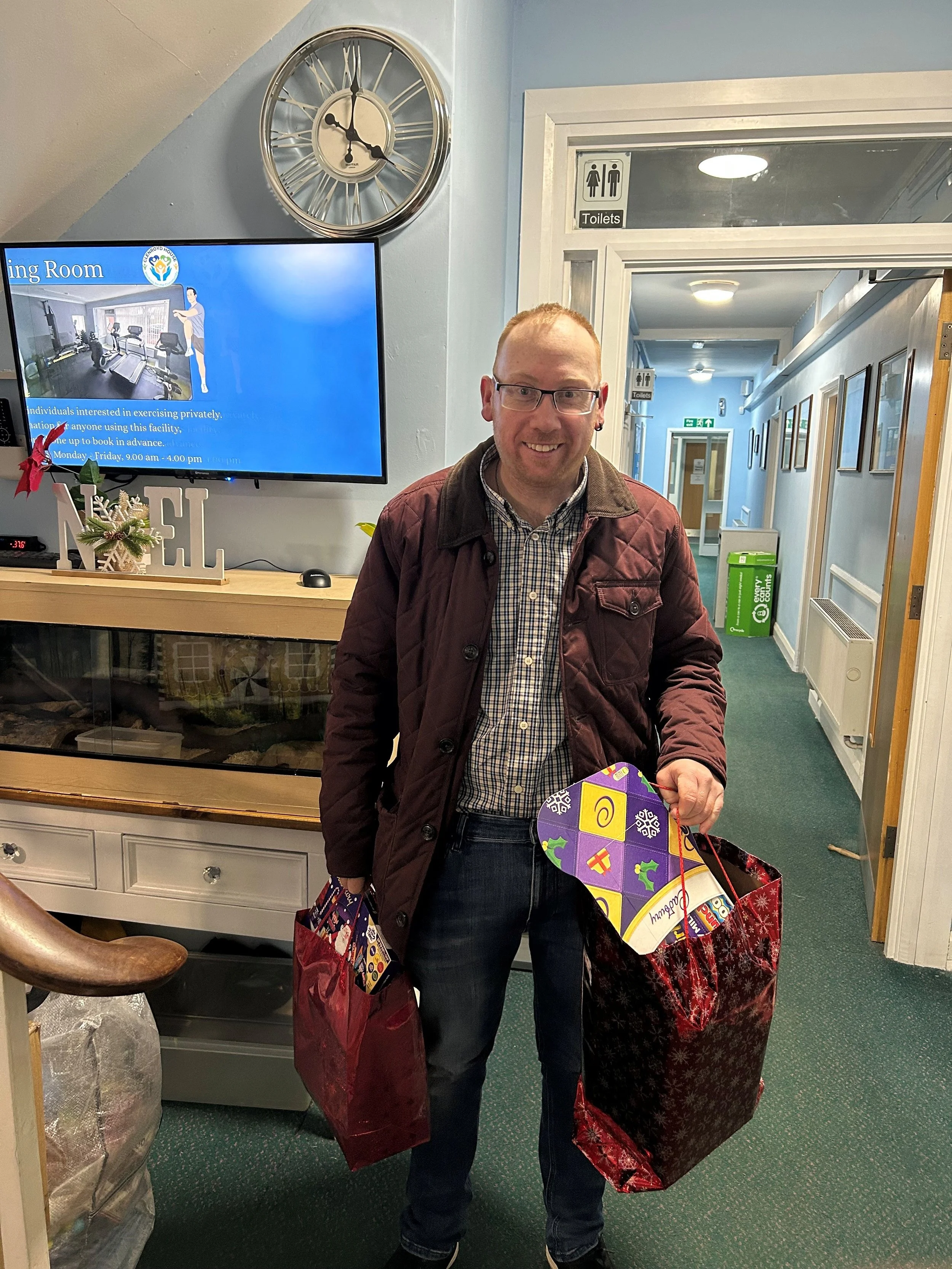 A smiling man holding large red shopping bags and a holiday gift box in an indoor setting with a hallway in the background.
