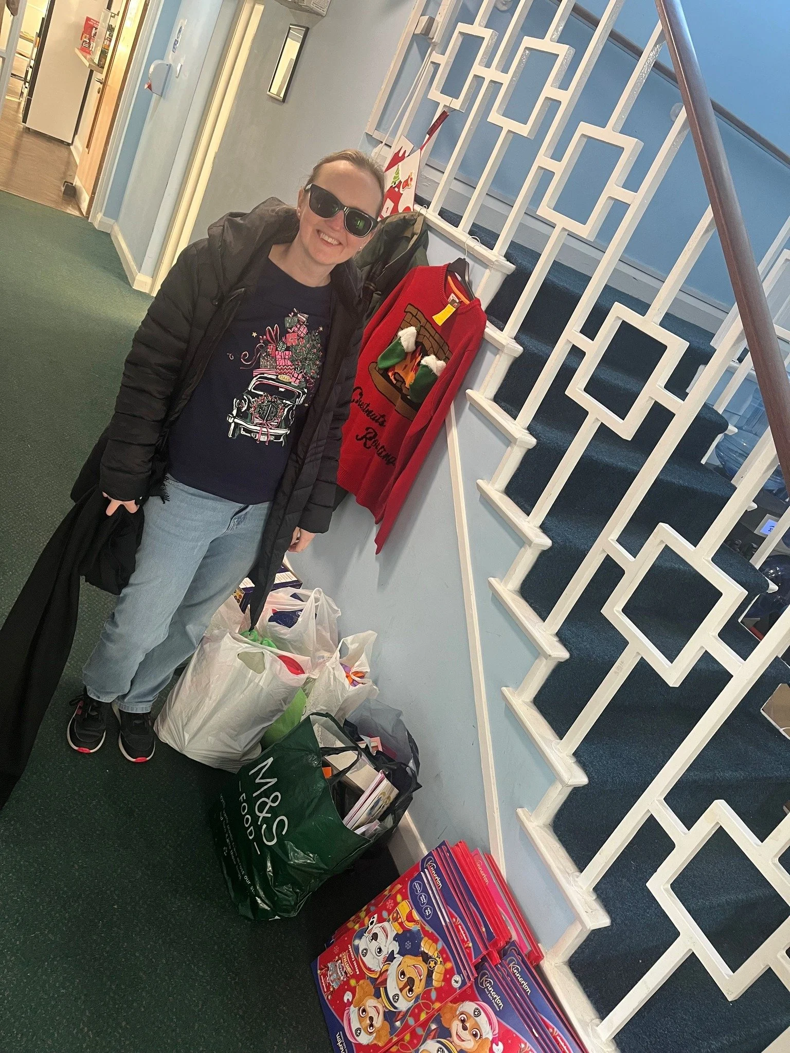 A woman smiling while standing on a green carpeted floor, surrounded by shopping bags, near a staircase decorated with Christmas stockings and a holiday sweater hanging on the railing.