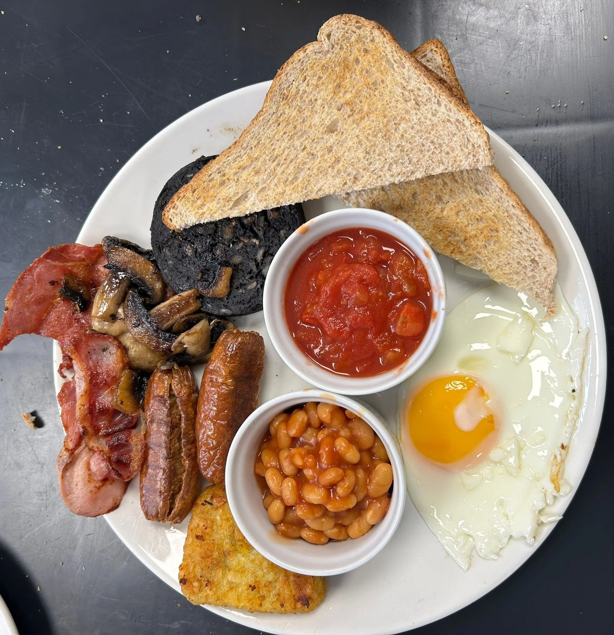 Full English breakfast on a white plate, includes fried egg, baked beans, grilled mushrooms, bacon, sausage, black pudding, toast, and tomato sauce.