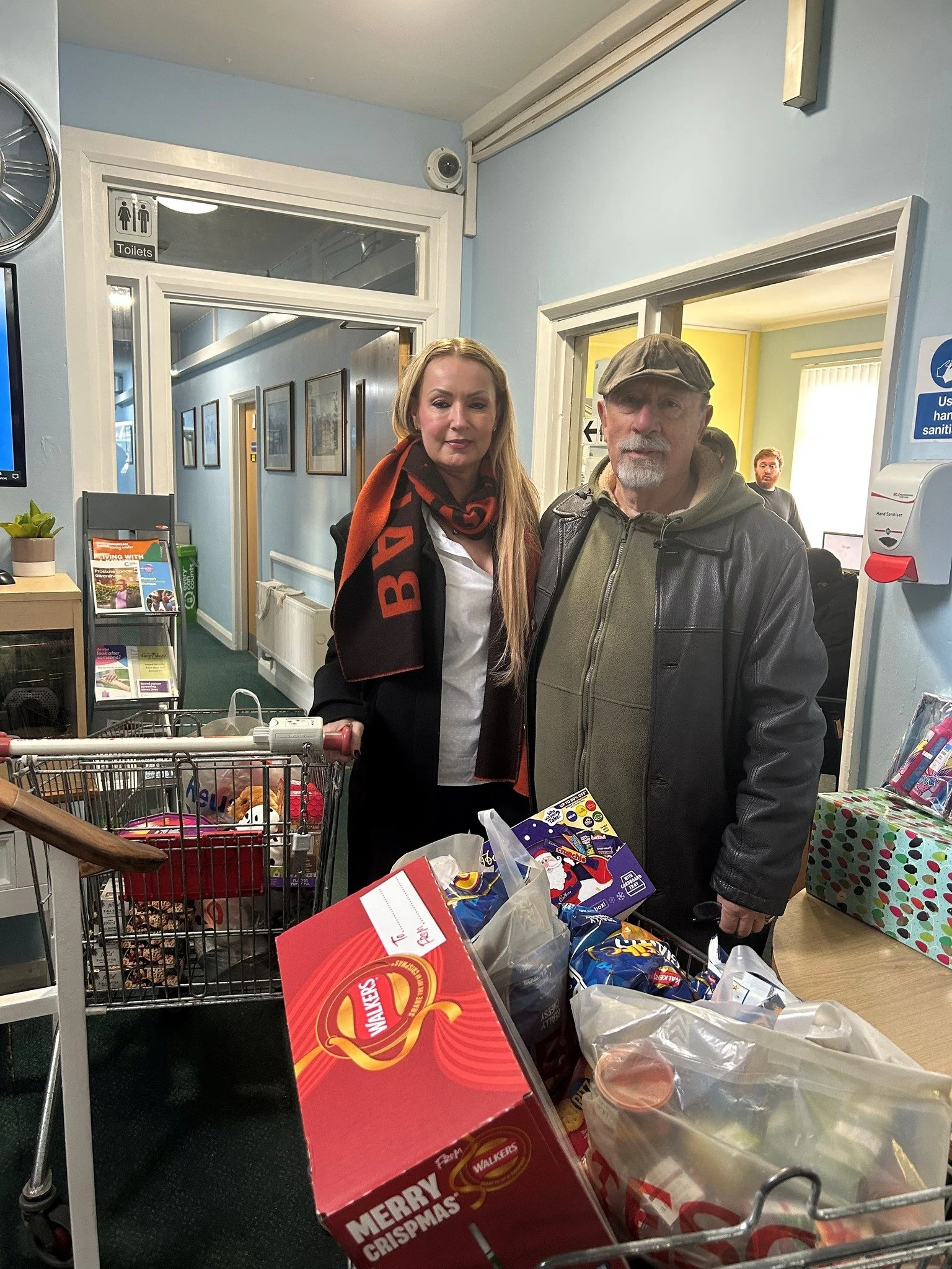 Two people, a woman and a man, standing in a room with grocery shopping carts filled with boxes and snacks, inside a building with blue walls and framed pictures.
