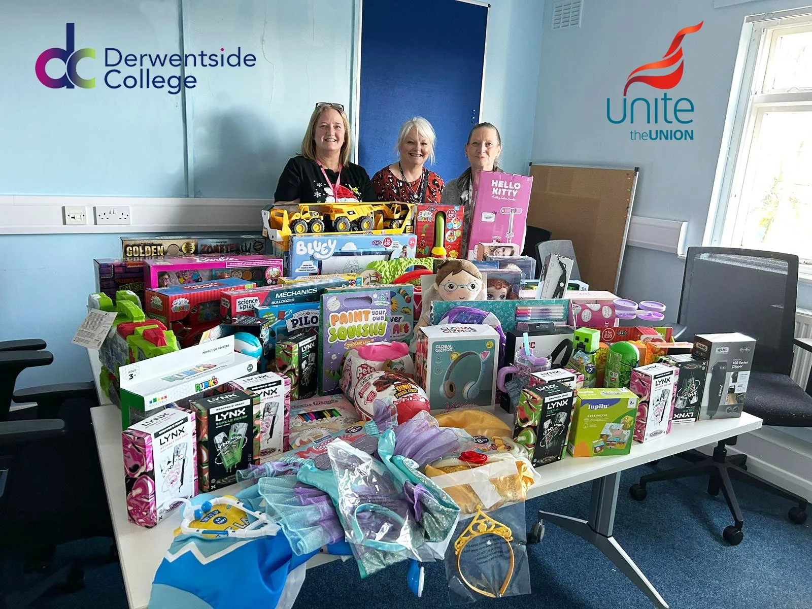 Group of three women standing behind a table filled with toys and gifts in a room with blue walls and windows, featuring Derwentside College and Unite the Union logos.