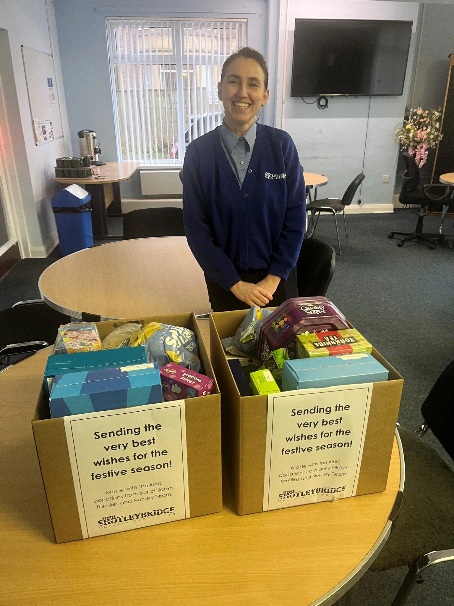 A woman standing behind two boxes filled with donations, inside a room with tables, chairs, a window, and a TV. The donation boxes have signs that read "Sending the very best wishes for the festive season!" from a nursery school.