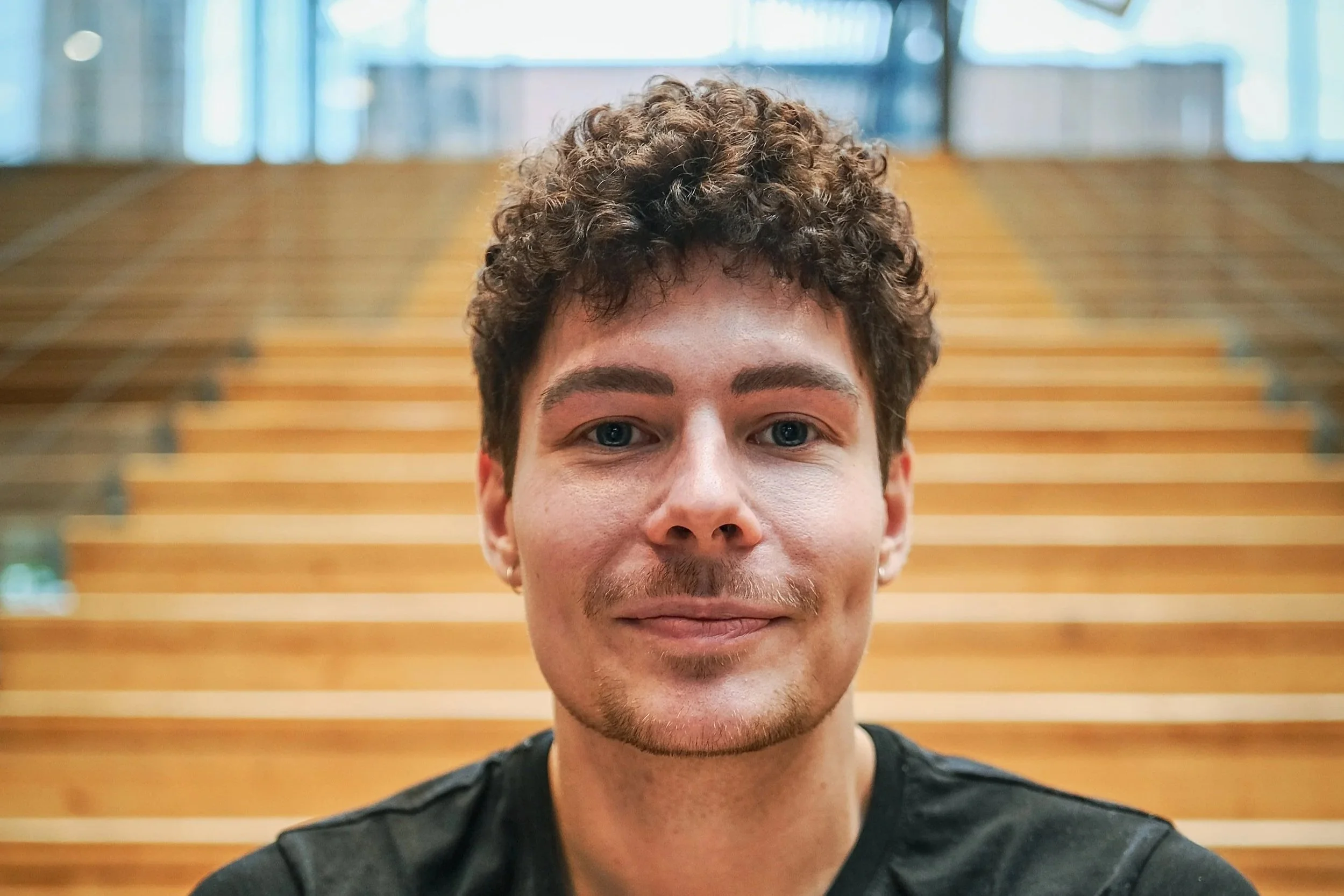 Young man with brown hair and fair skin wearing a black shirt against a purple and pink gradient background.