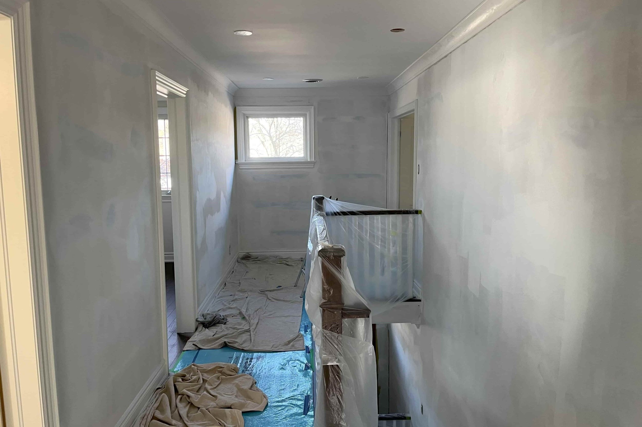 Interior hallway under renovation with plastic sheets and protective coverings on the railing and floor, unfinished walls, and a window at the end.