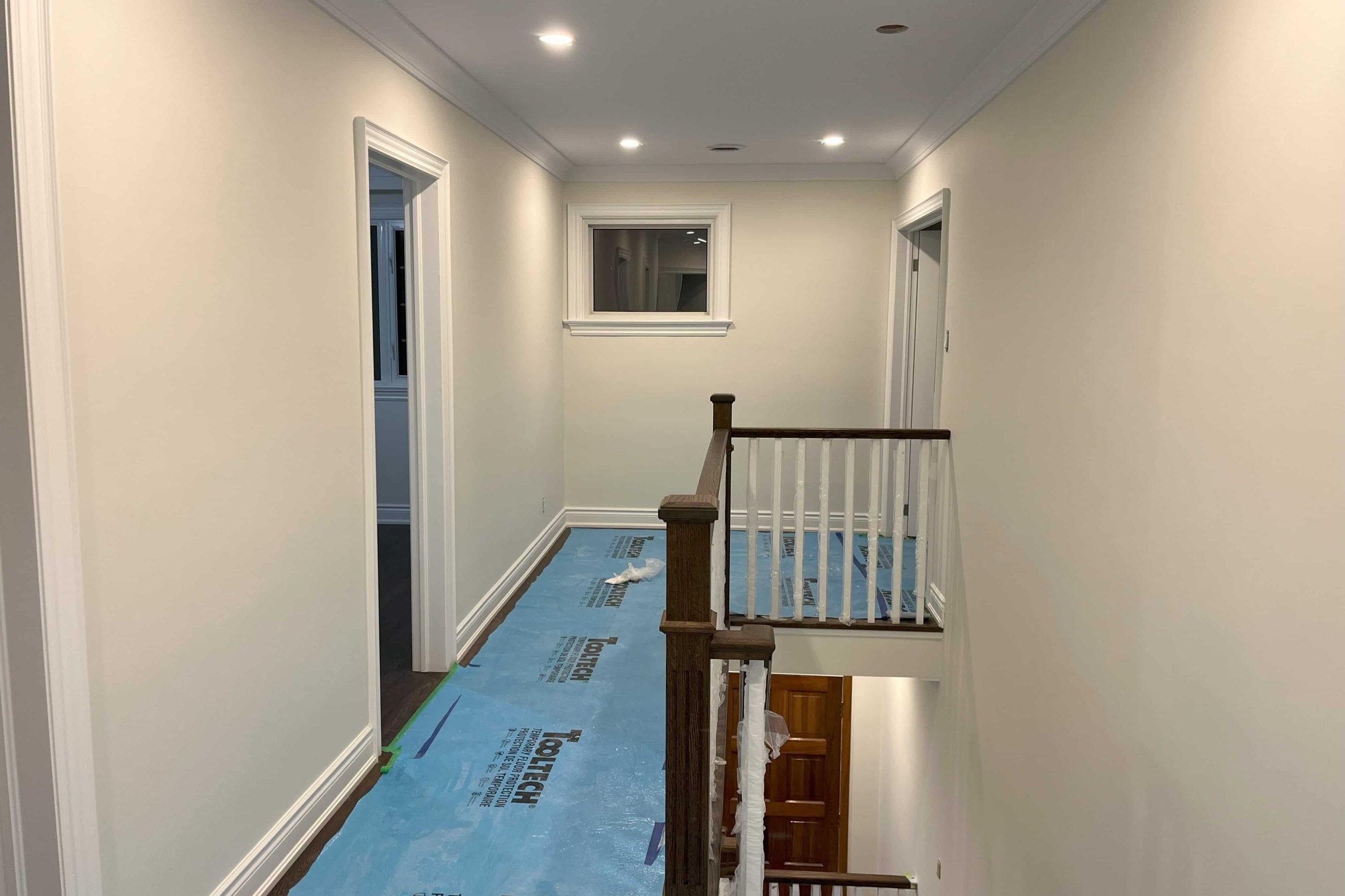 Interior hallway with white walls, recessed lighting, a wooden railing, and blue protective floor covering.