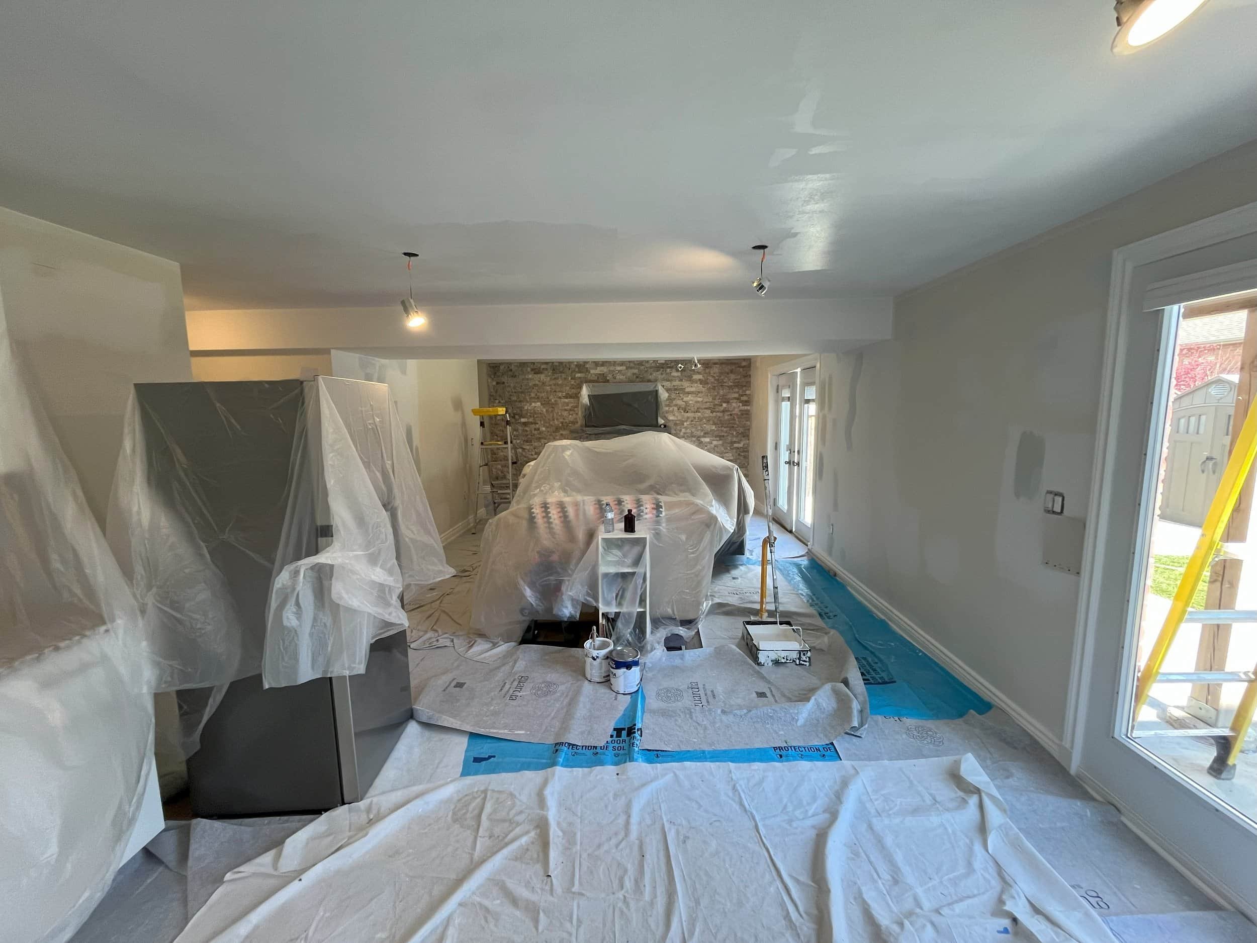 Living room under renovation with plastic coverings, paint supplies, ladder, and partially painted walls.