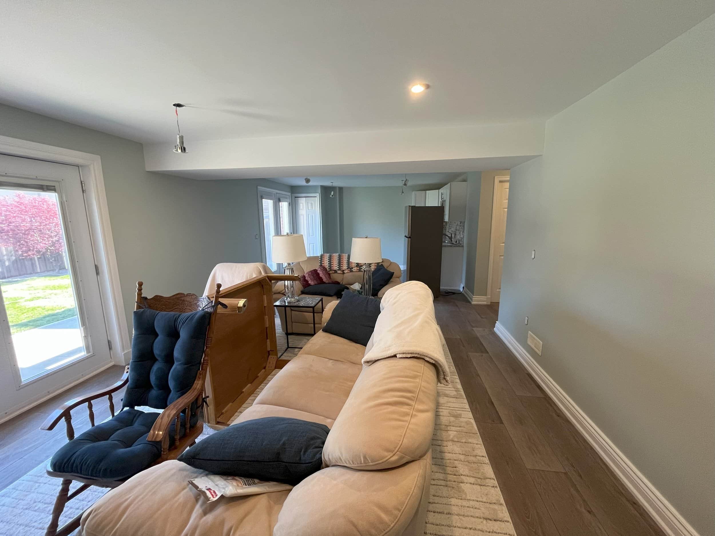 Living room with beige couch, wooden rocking chair with dark blue cushion, side tables, lamps, and sliding glass door leading outside, with an open kitchen in the background.