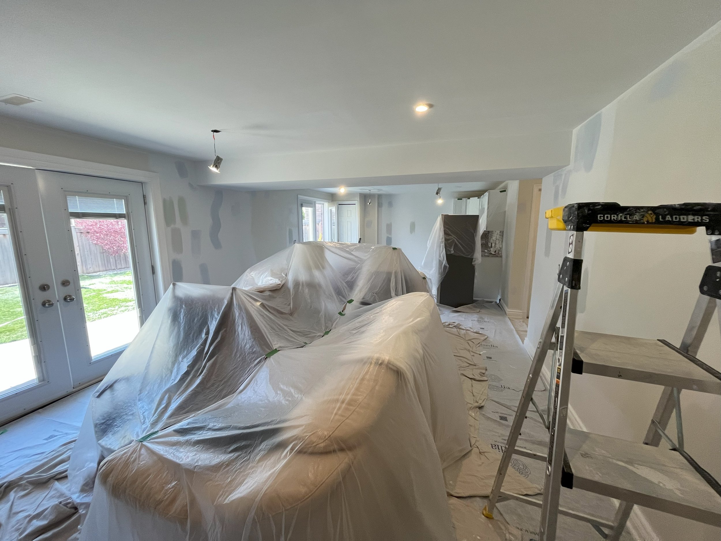 Living room under renovation with furniture covered in plastic, ladder, and drywall patches on walls. All set to start painting by expert interior painters.