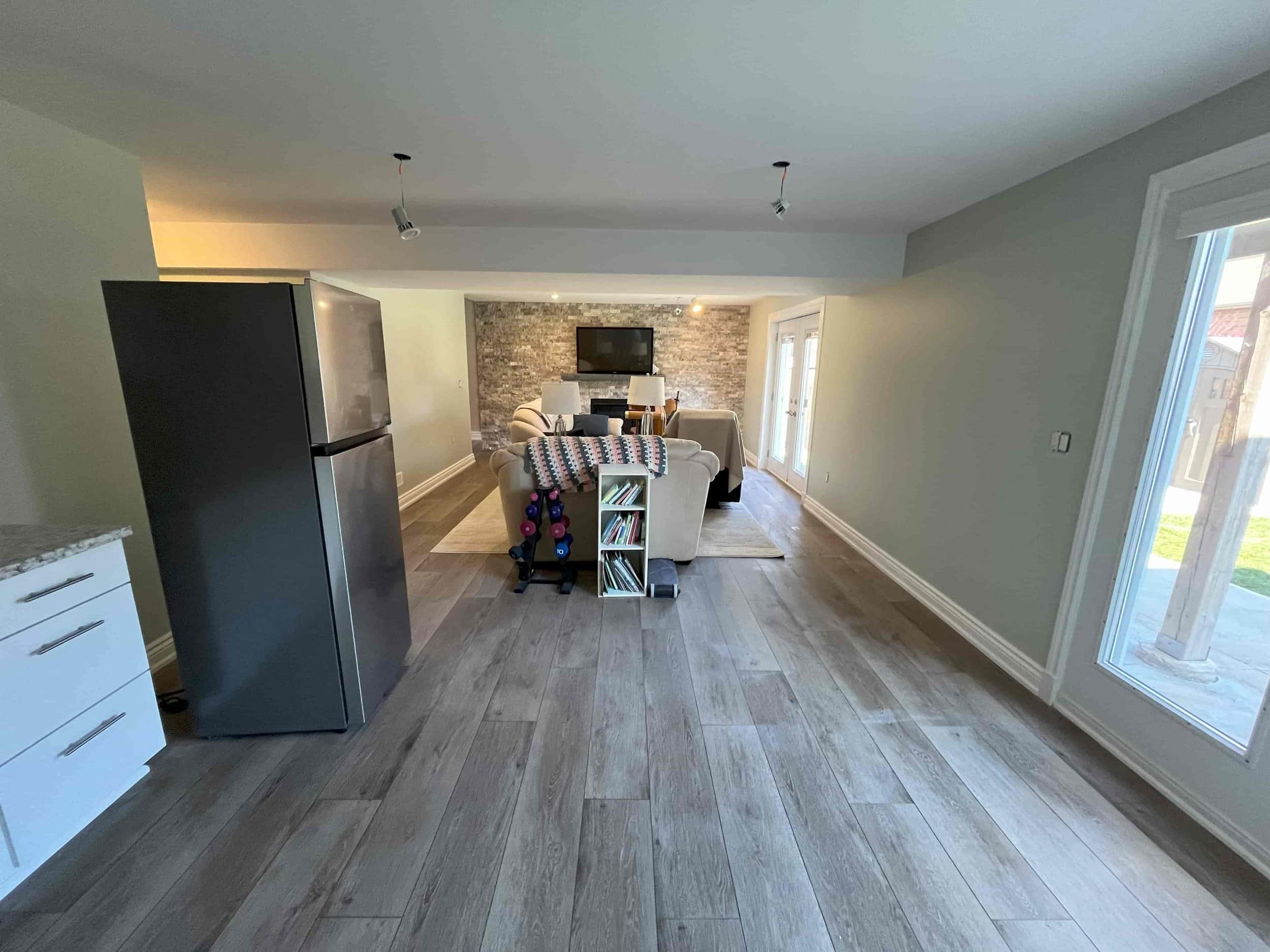 Open-concept living room with hardwood floors, a gray brick accent wall, a wall-mounted TV, cream-colored sofas, a small bookshelf, and French doors leading outside.