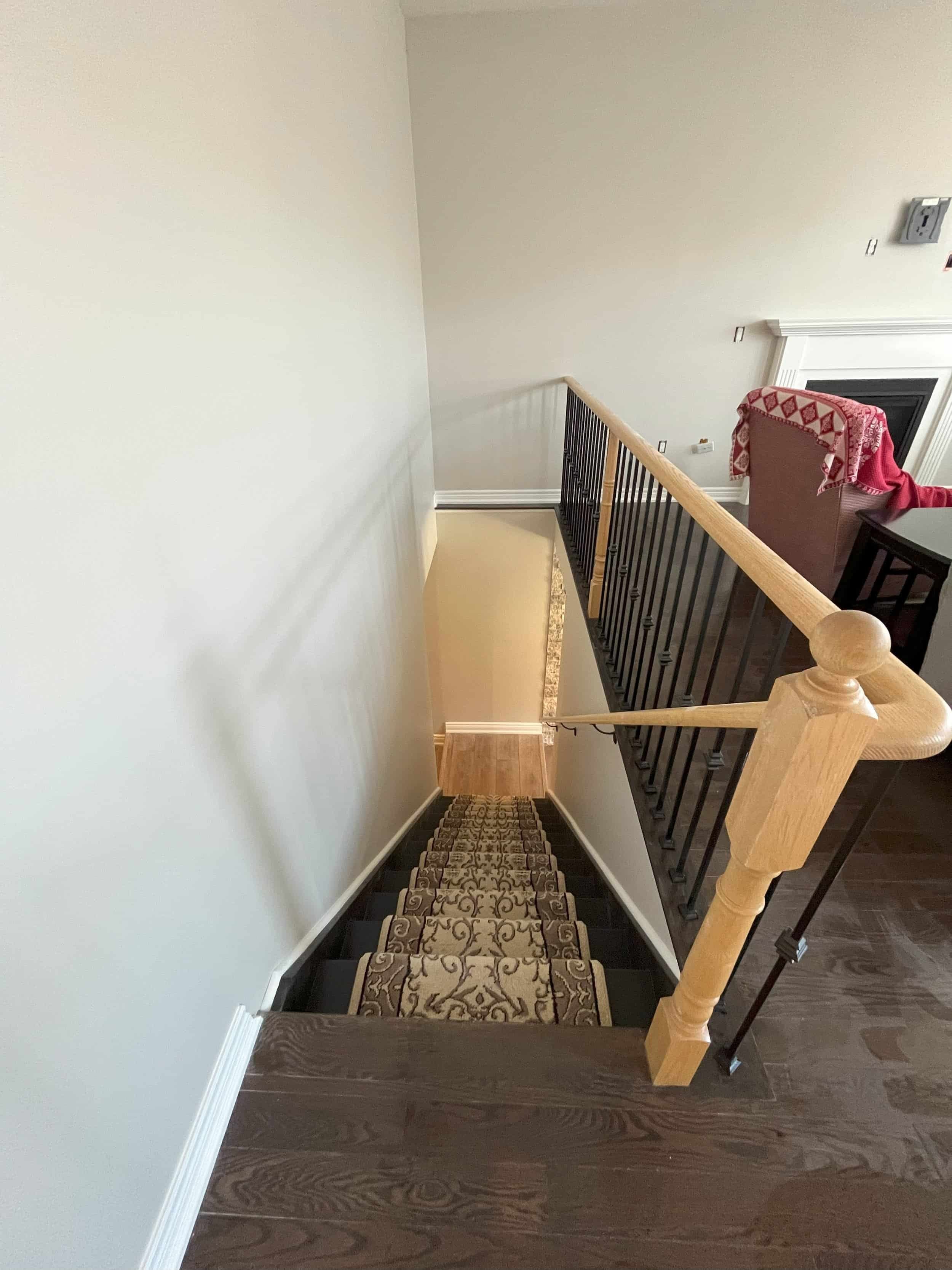 Interior view of a staircase with dark carpeted steps featuring a decorative pattern. The staircase has a wooden handrail and black metal balusters, descending to a landing with wooden flooring. To the right, part of a dining area or living space is