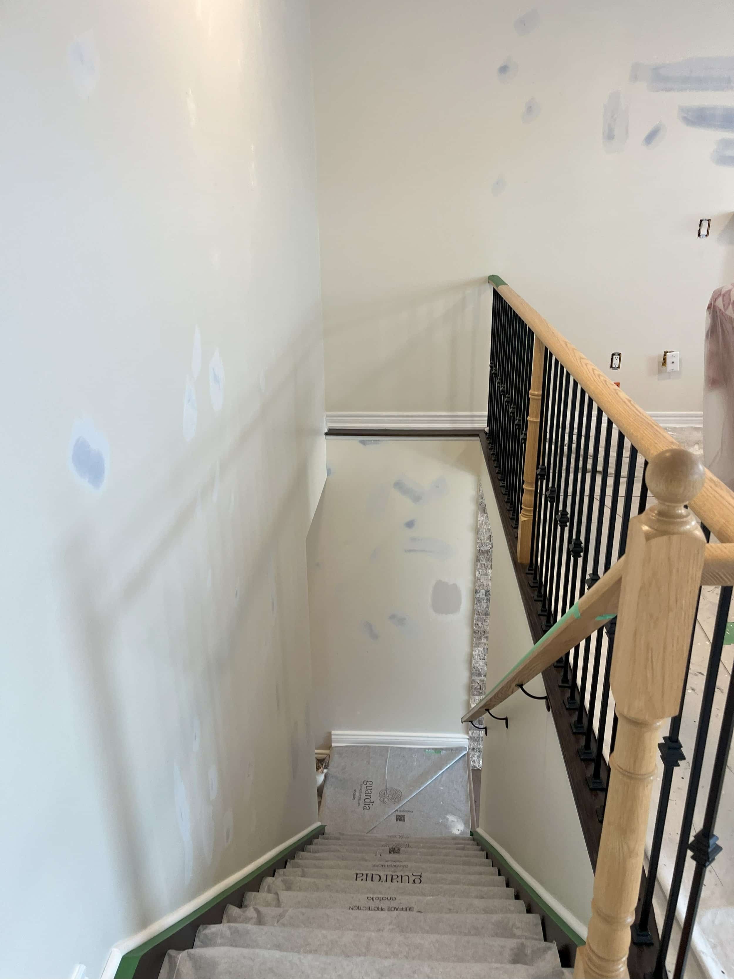 Interior staircase under construction, with drywall patches on the walls, stair treads covered in protective paper, and unfinished electrical outlets.