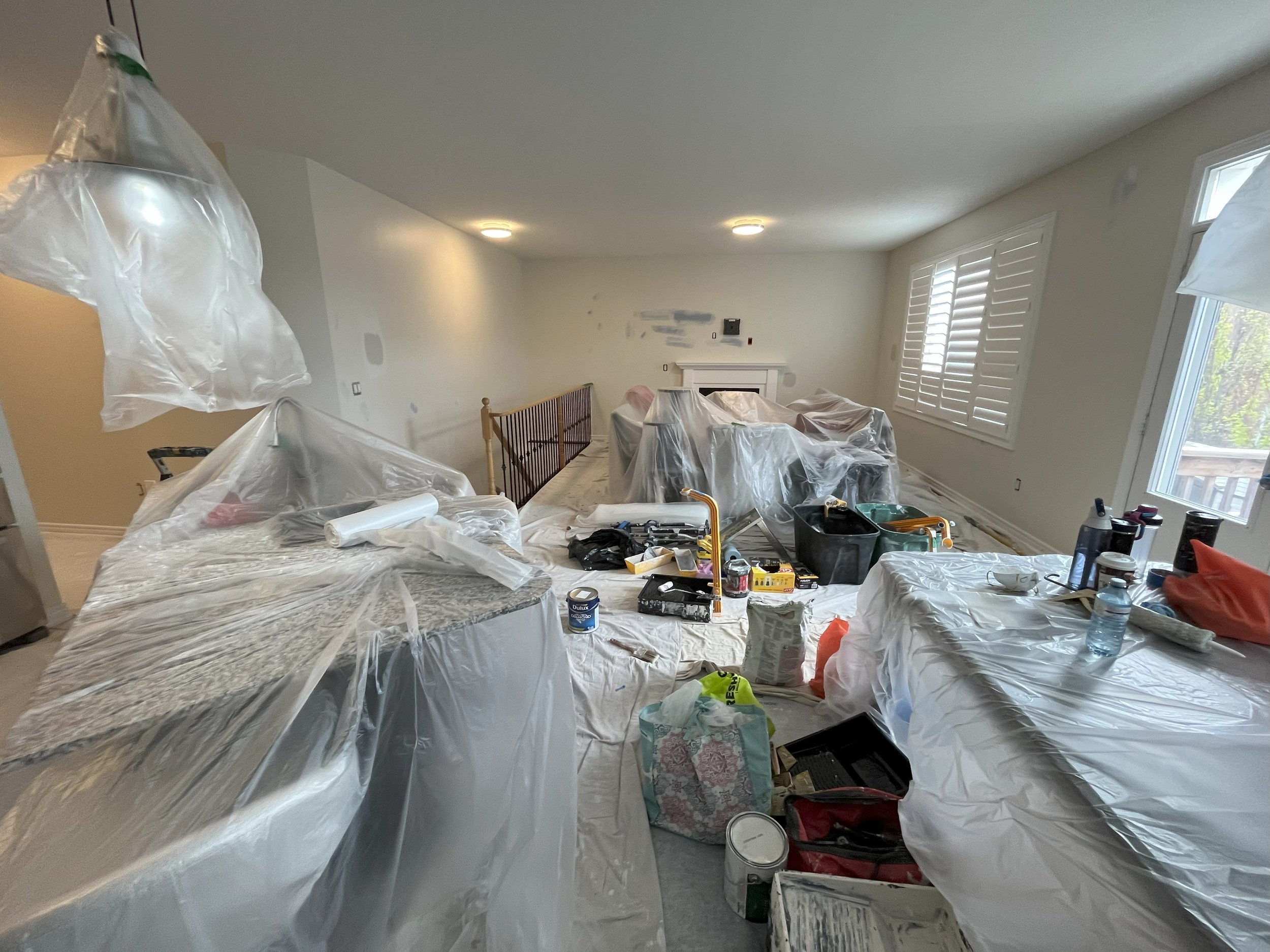 Living room under renovation with furniture covered in plastic, painting supplies, and various tools. Getting ready to be painted by expert interior painters.