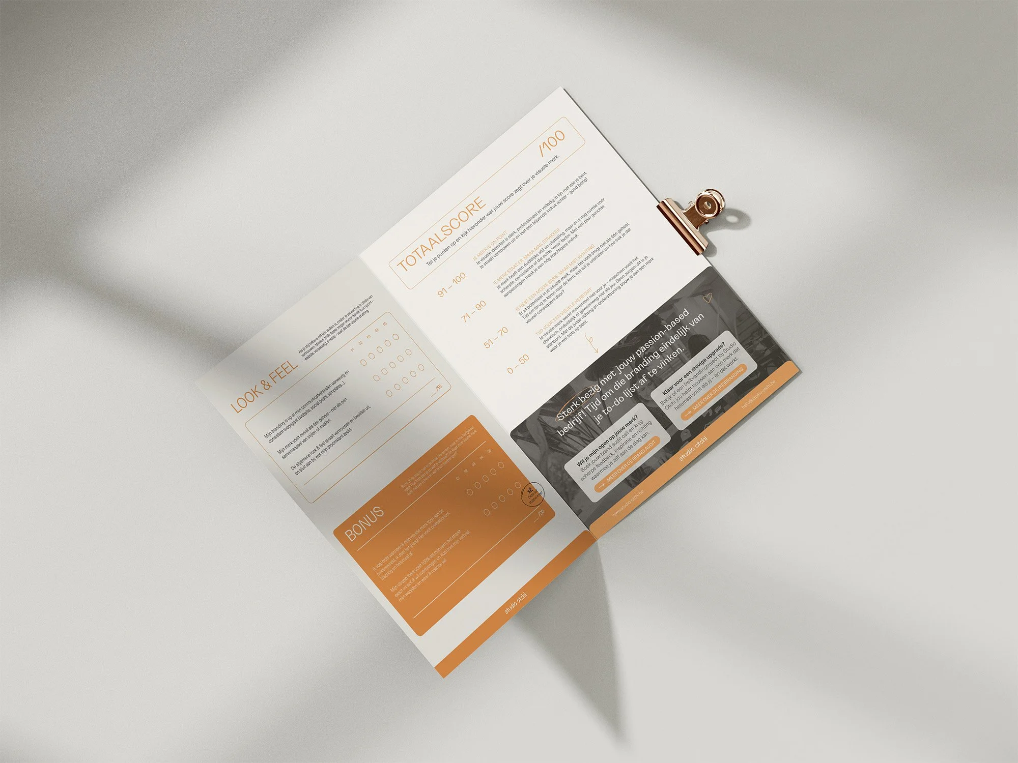 Open brochure with orange accents, containing sections labeled 'LOOK & FEEL,' 'TOTAL SCORE,' and a social media post, clipped with a gold clip on a plain white background.