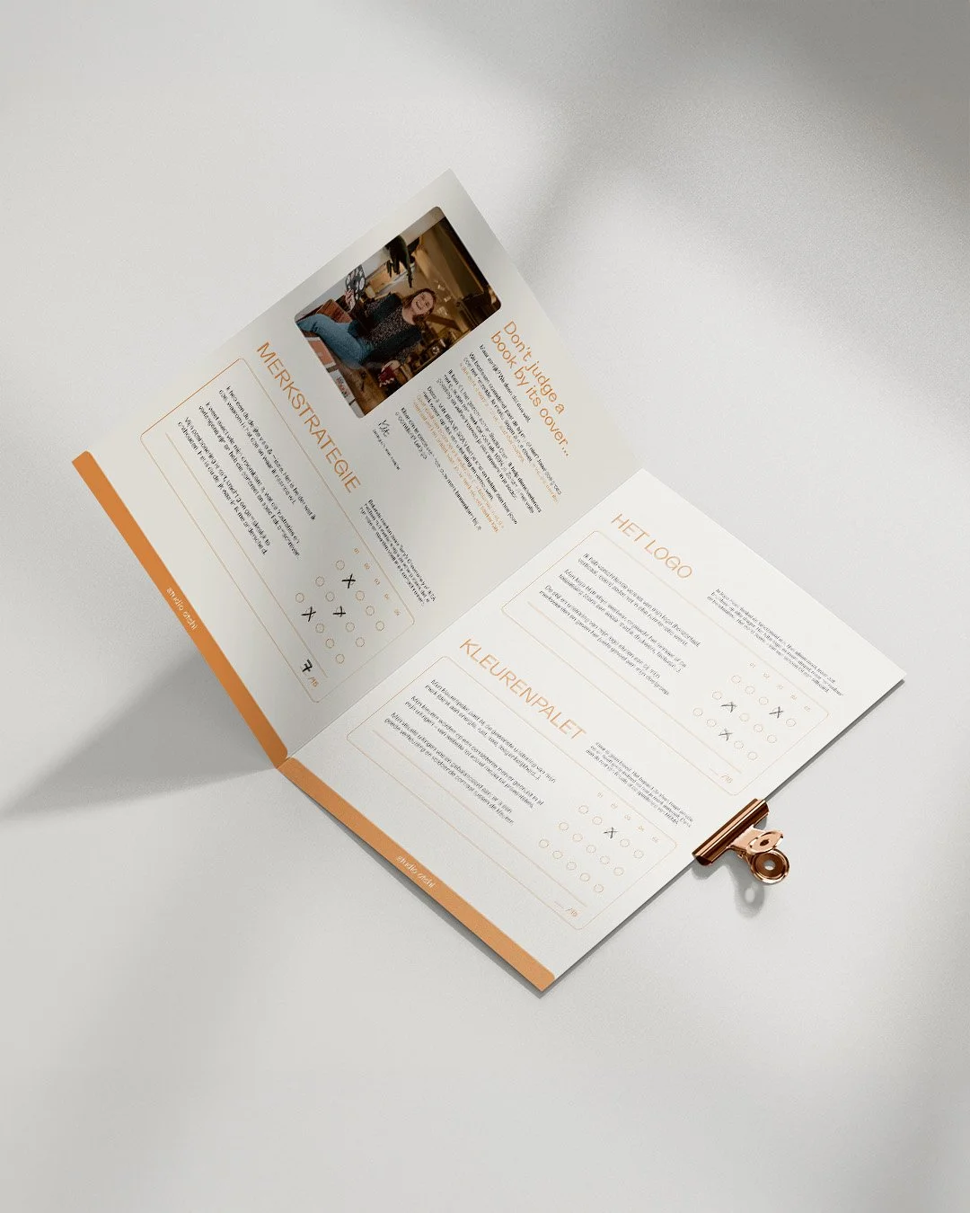 Open booklet with orange accents, containing text and diagrams on the left and right pages, clipped with a rose gold metal clip, on a white surface.