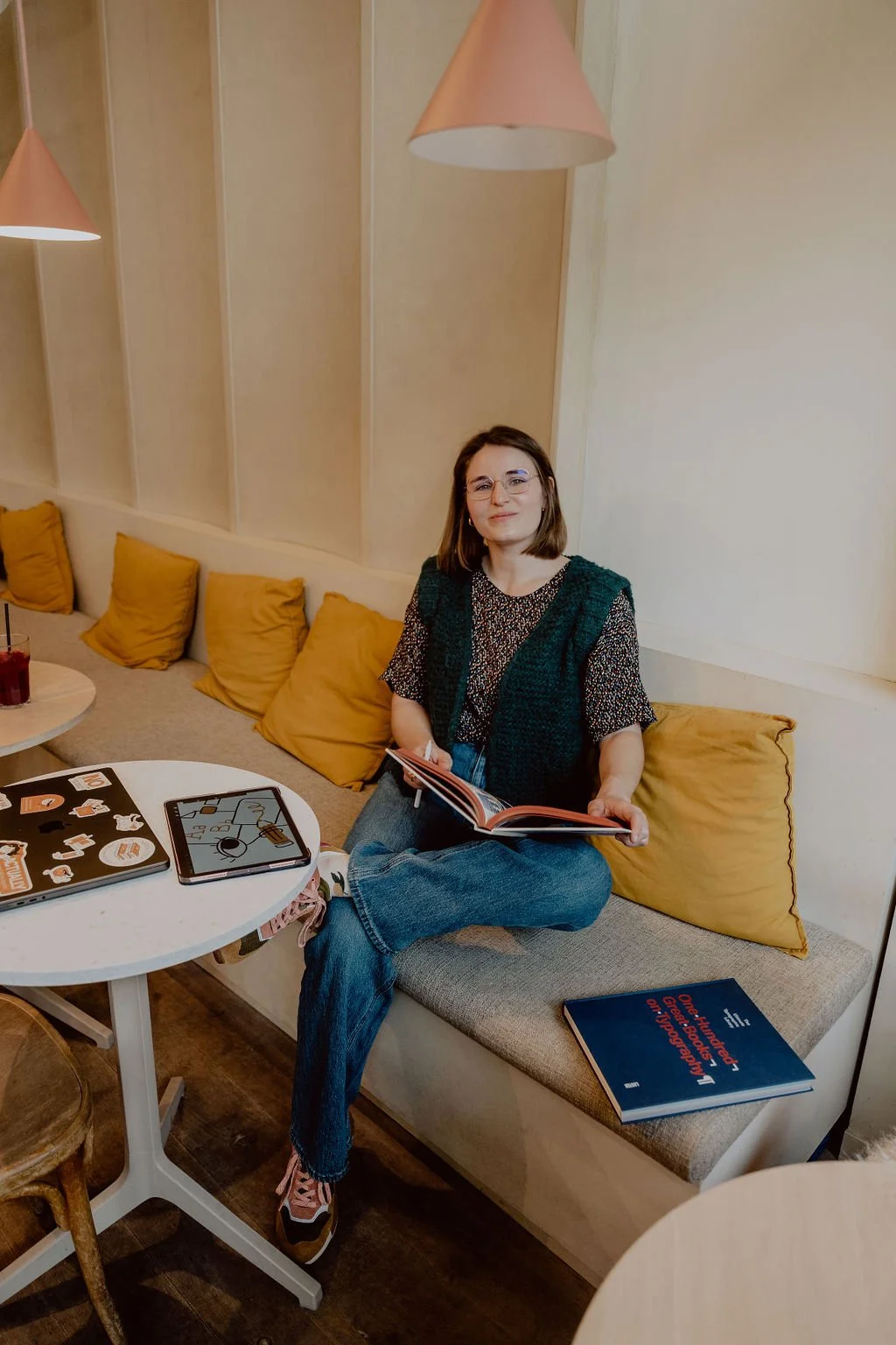 A young woman with shoulder-length brown hair, glasses, and a small smile, sitting on a cushioned bench in a cozy cafe. She is holding an open book in her hands, wearing a patterned shirt, a knitted vest, and jeans. There are four yellow pillows behind her, a small round table with a laptop covered in stickers, a closed laptop, and a book on her lap. The cafe has warm lighting with pink pendant lights hanging from the ceiling.