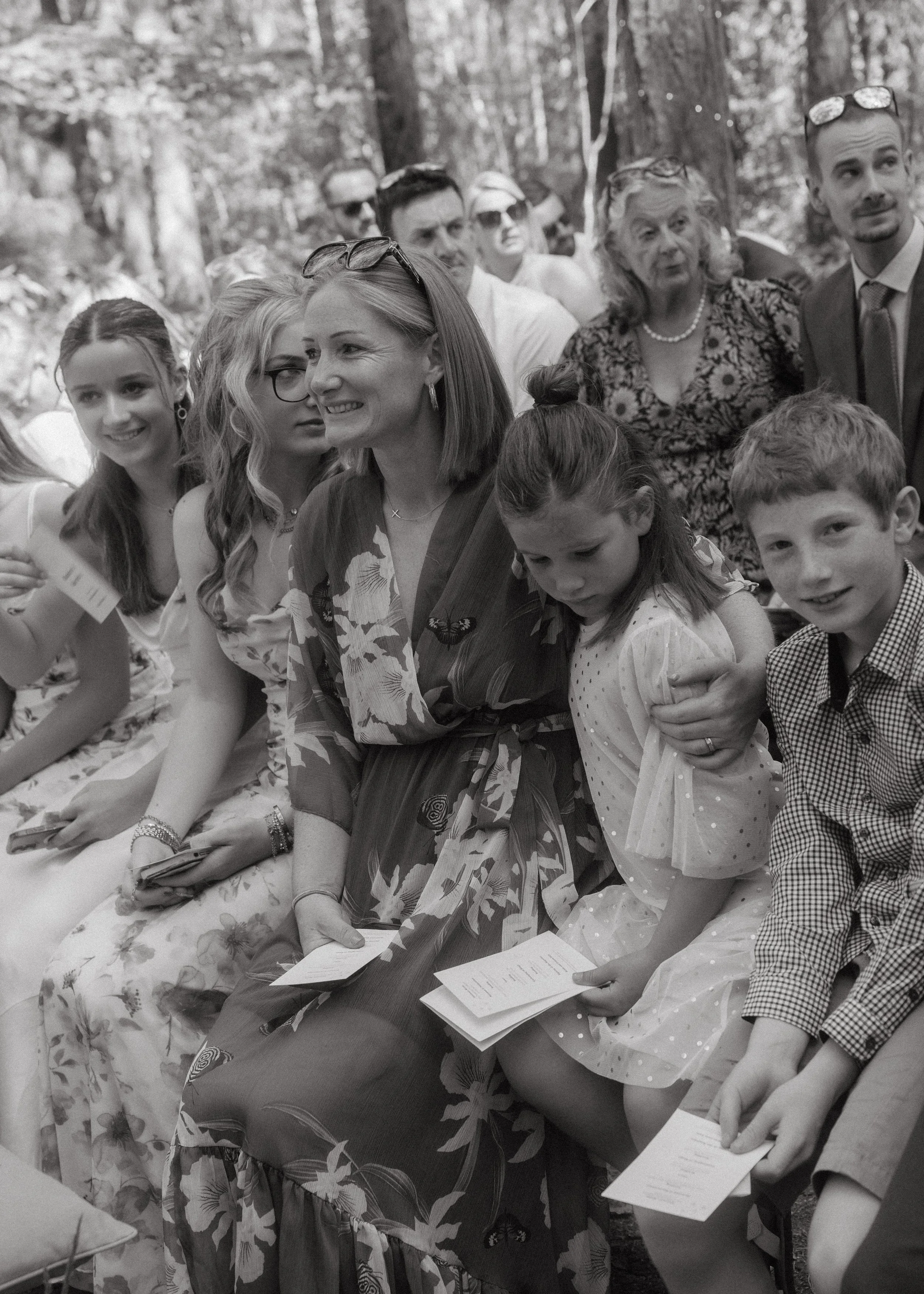 A group of people sitting outdoors, dressed formally, appearing to be at a social event or gathering.
