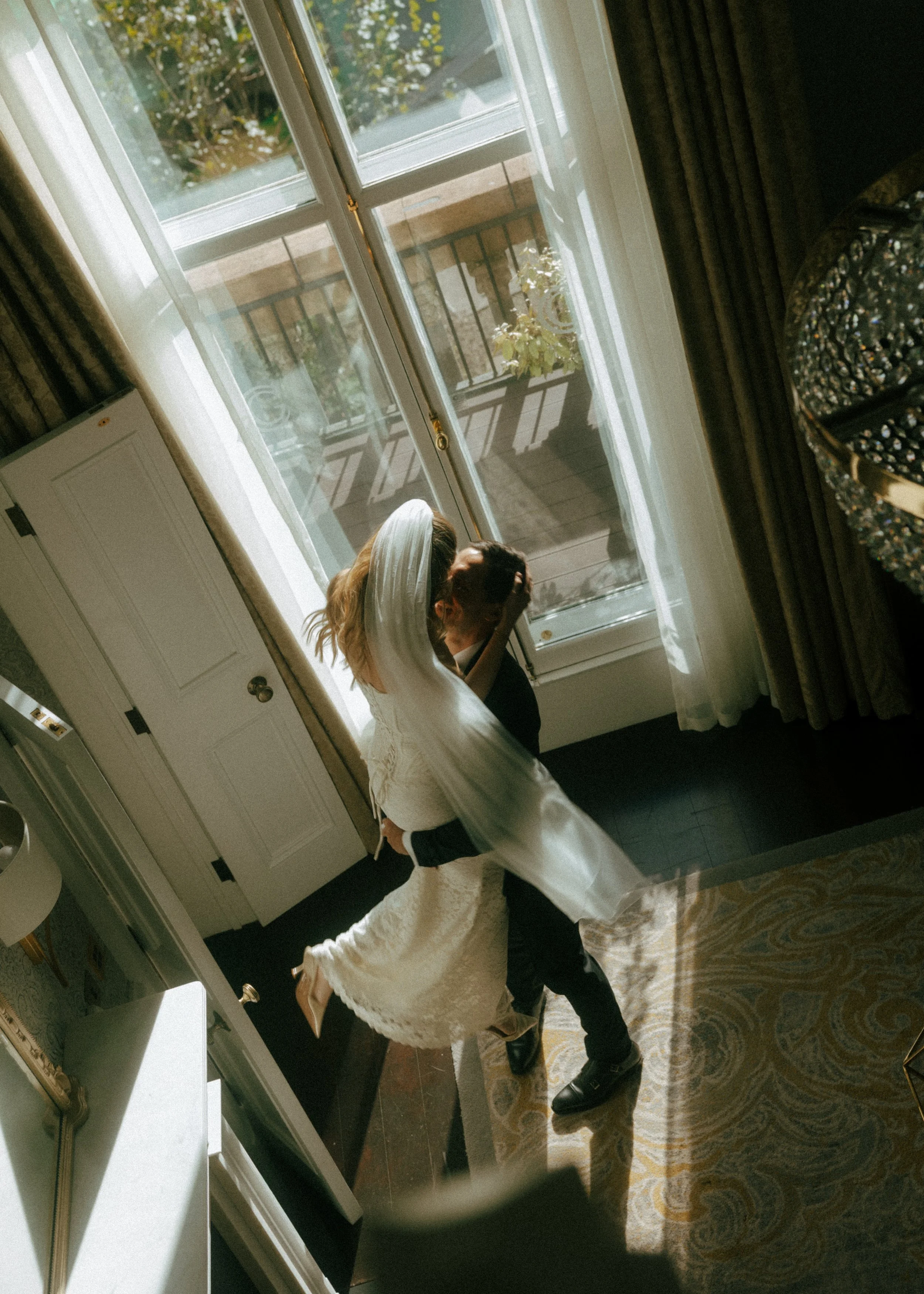 A bride in a white wedding dress and a groom in a black suit kissing inside a room near glass doors that lead to a balcony.