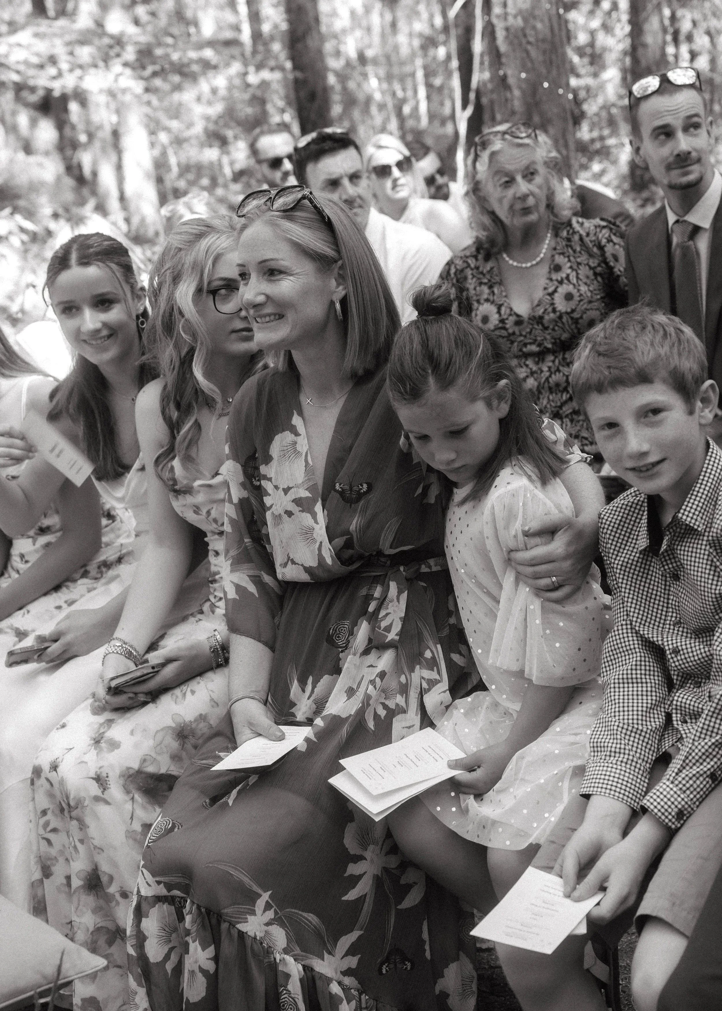 A group of people sitting outdoors, dressed formally, appearing to be at a social event or gathering.