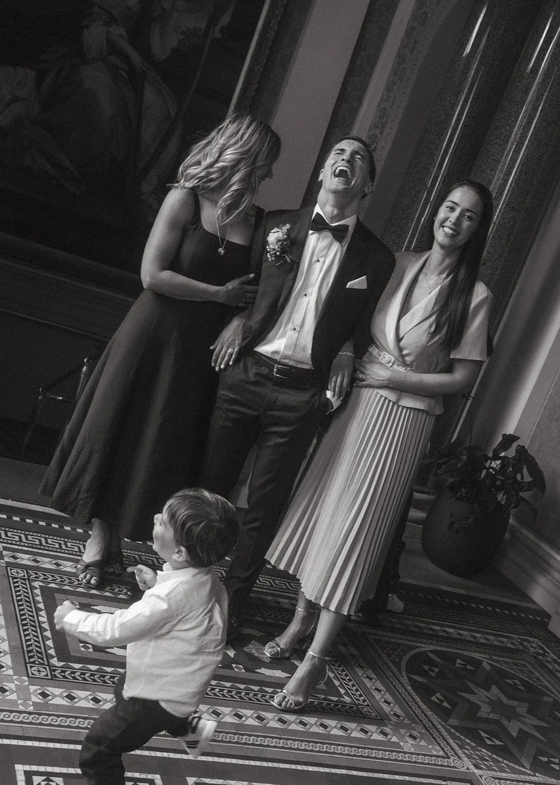 Three adults and a child at a formal event. The man is in a tuxedo and the two women are in elegant dresses. The child is running in front of them.