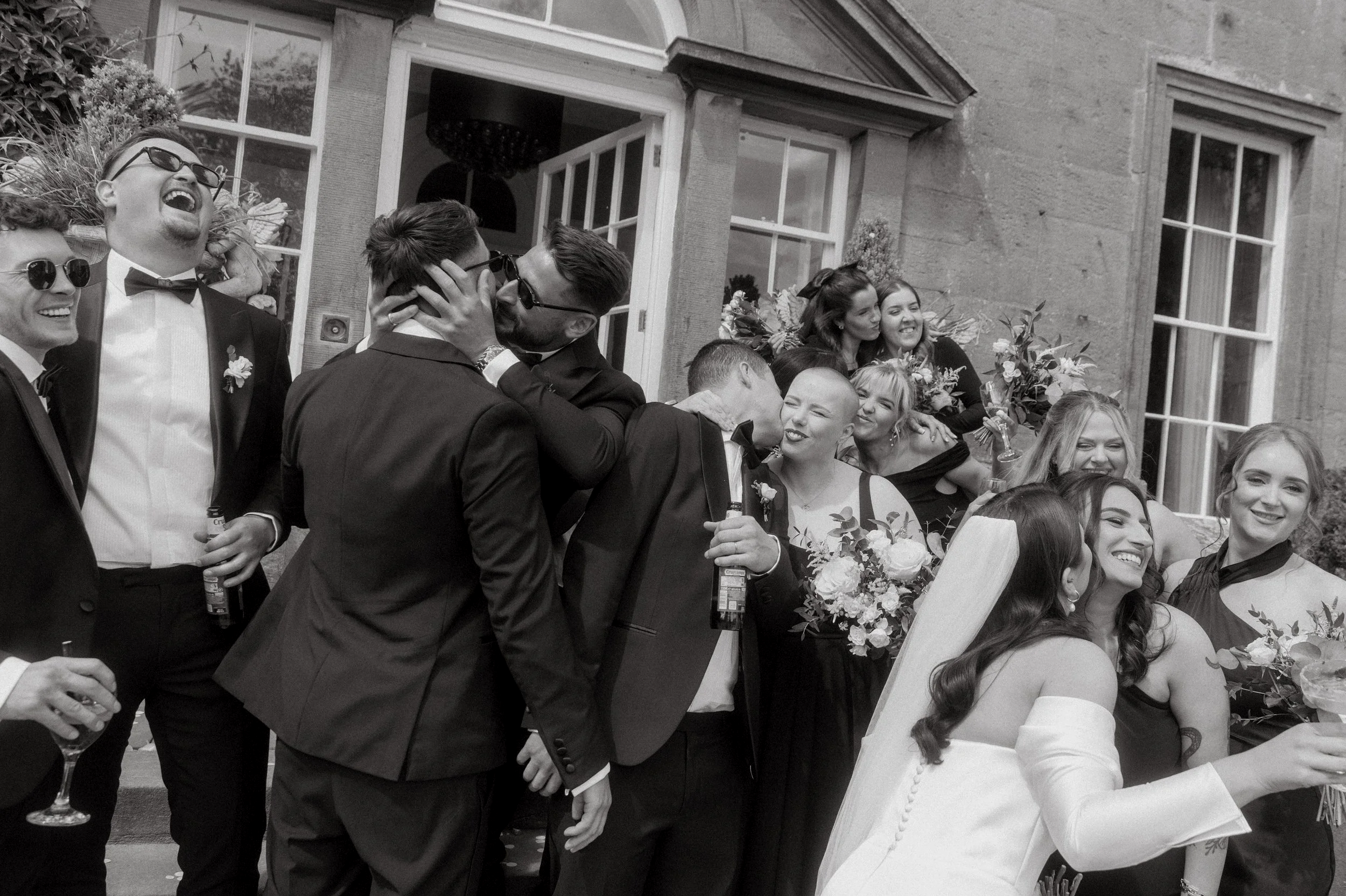 A group of people at a wedding party celebrating outside a building, some wearing tuxedos and holding drinks, others in dresses, laughing and engaging with each other.