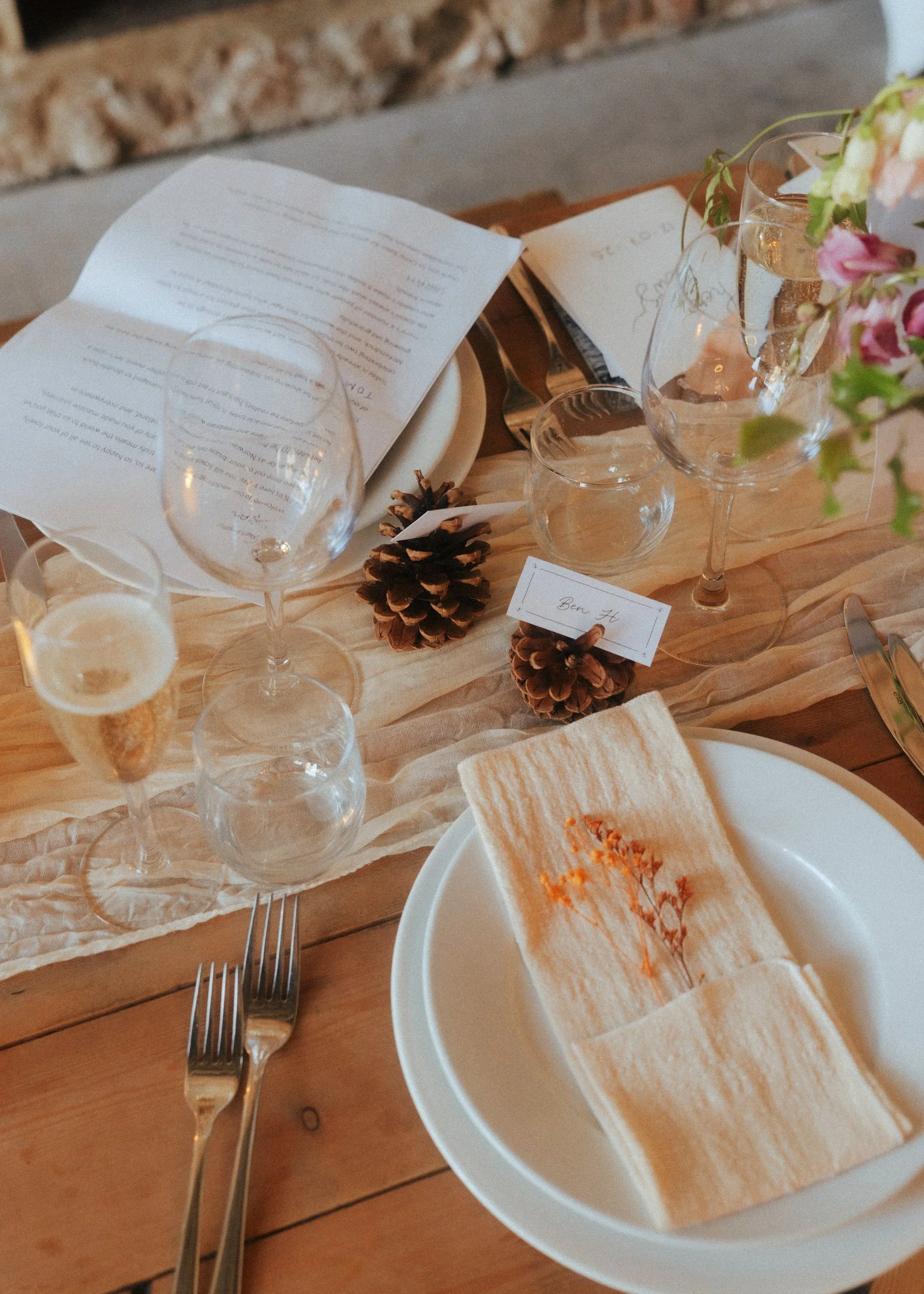 Table setting with wine glasses, a pinecone, an open menu, and a small card that says 'Ben H.' on a wooden table with a peach-colored table runner.
