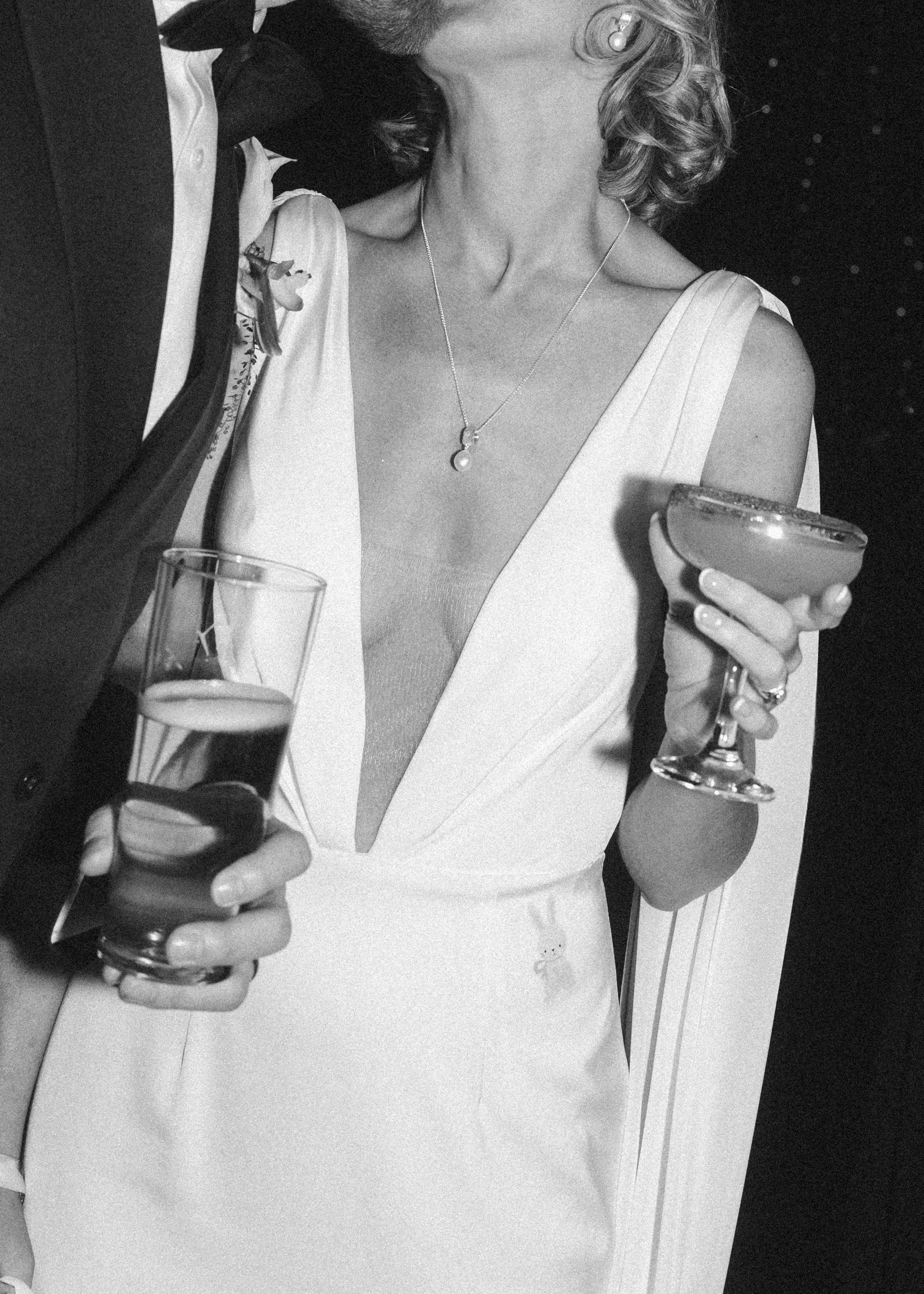 Black and white photo of a woman dressed in a white gown with a deep neckline, holding a glass of beverage, wearing jewelry, and standing next to a man in a dark tuxedo with a bow tie.