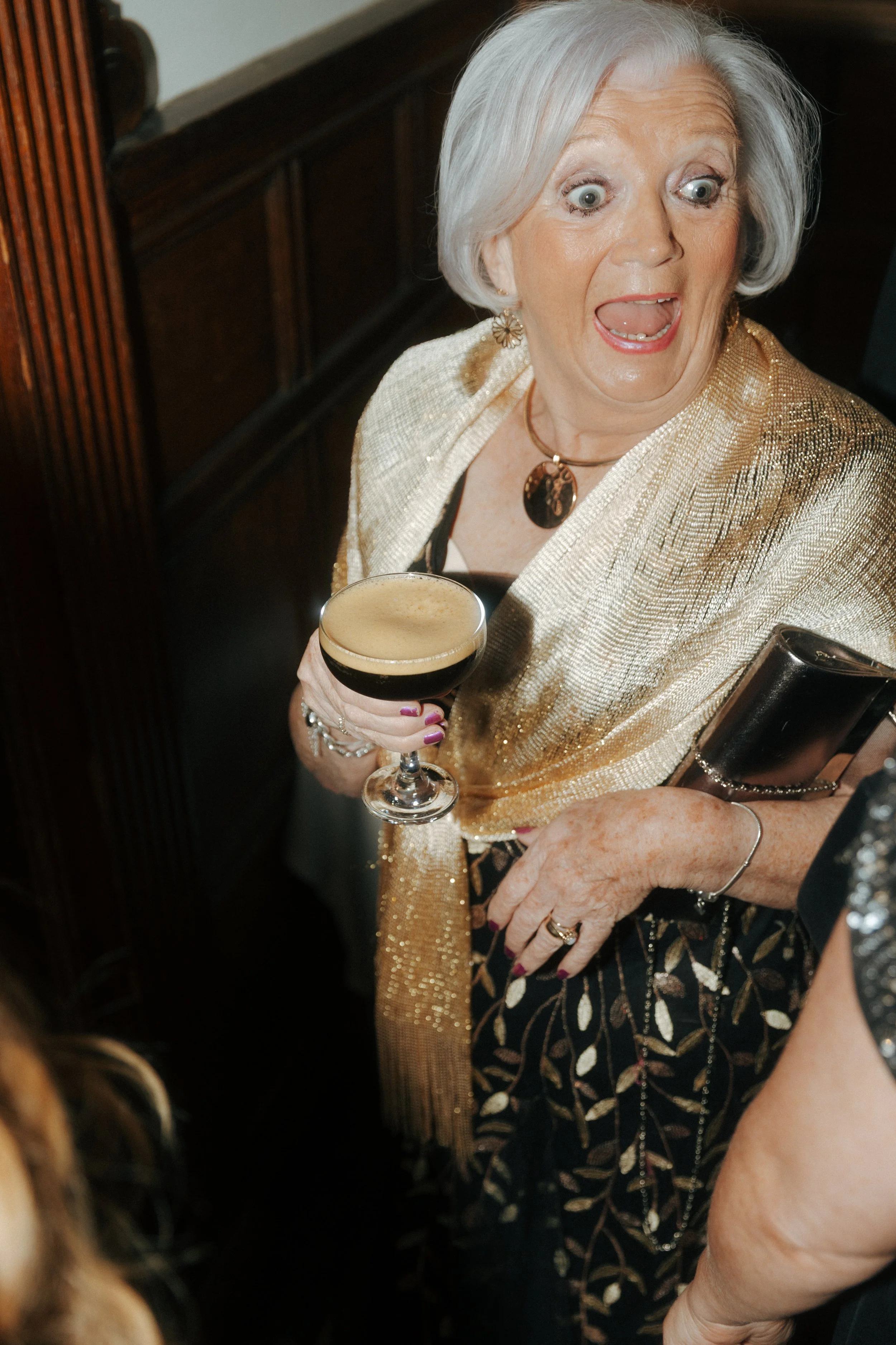 An elderly woman with gray hair, wearing a shiny gold outfit and jewelry, holding a glass of dark beer and looking surprised or excited.