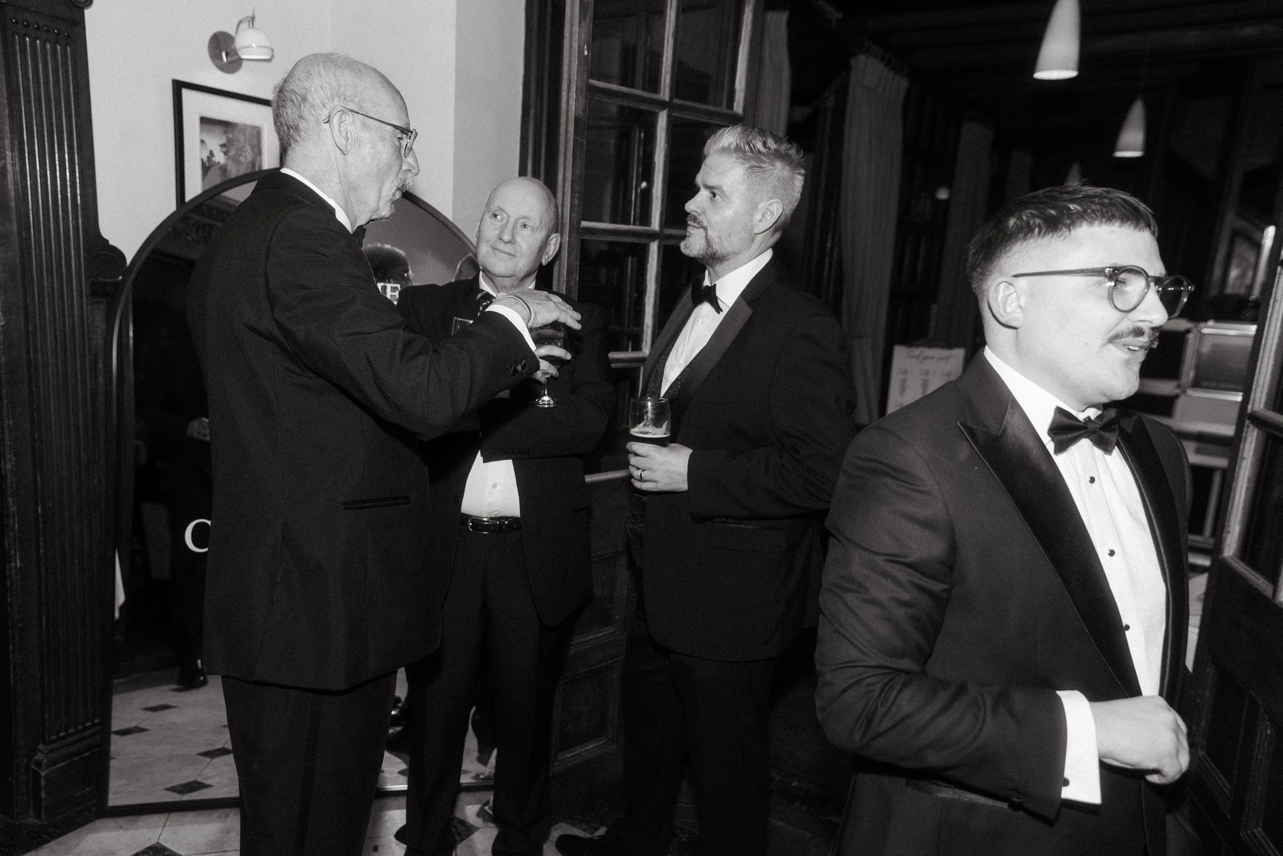 Four men in tuxedos at a social event, standing and talking in a room with wooden walls and windows, some holding drinks.