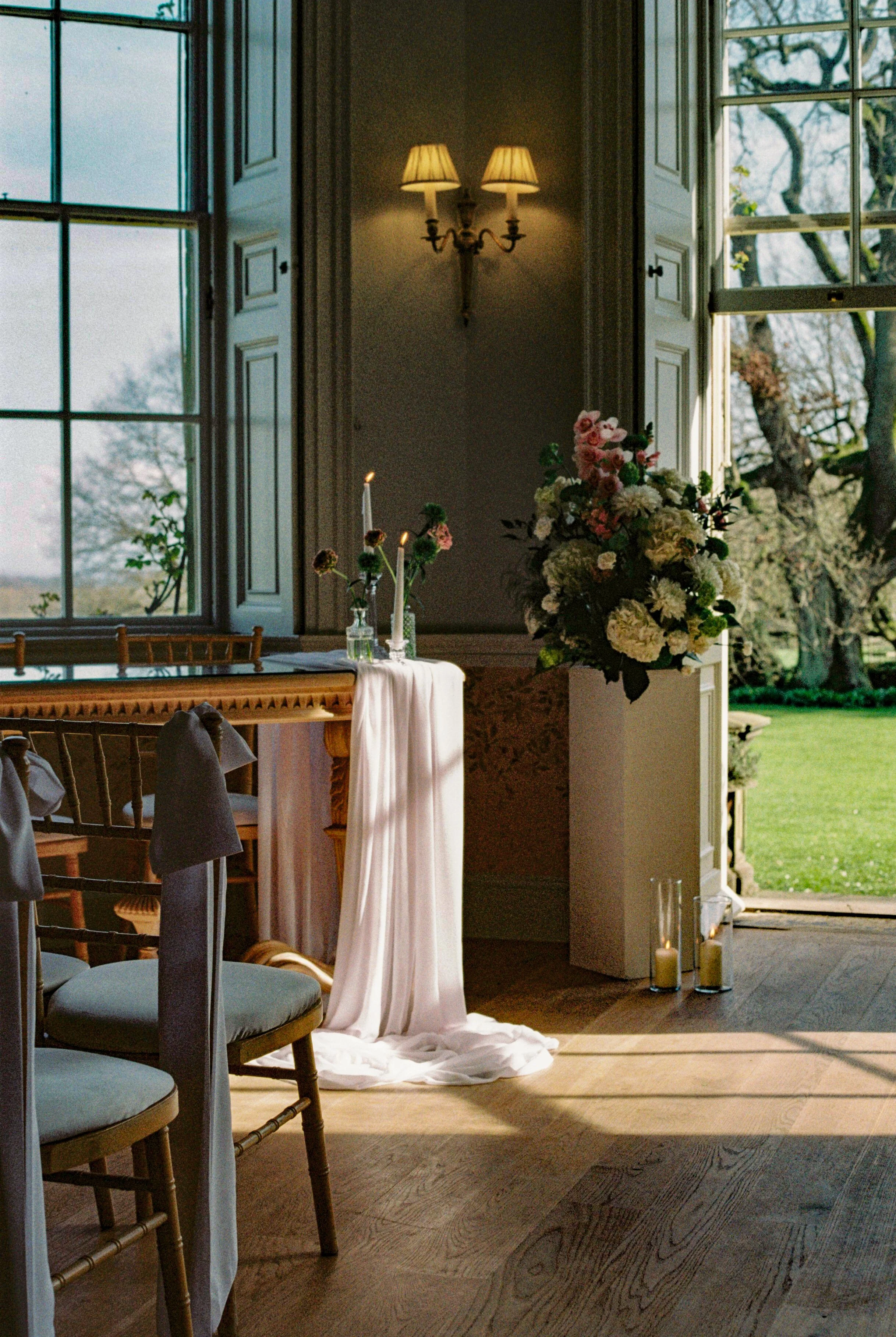 An indoor wedding or event setup with large open windows, a floral arrangement, lit candles, and a view of trees outside.