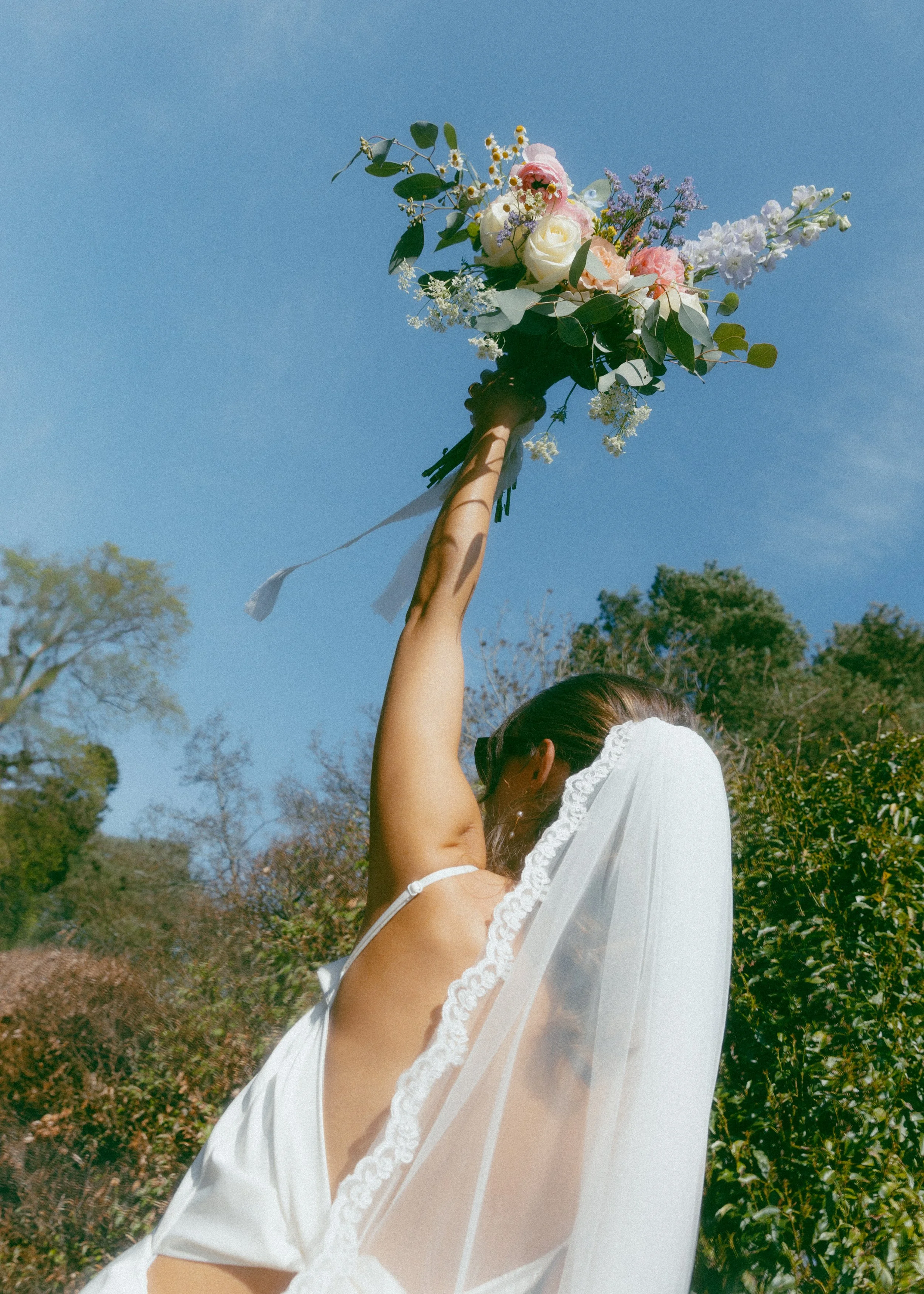 A woman wearing a white dress and veil holds a bouquet of flowers up to the bright blue sky. Trees with green leaves are in the background.
